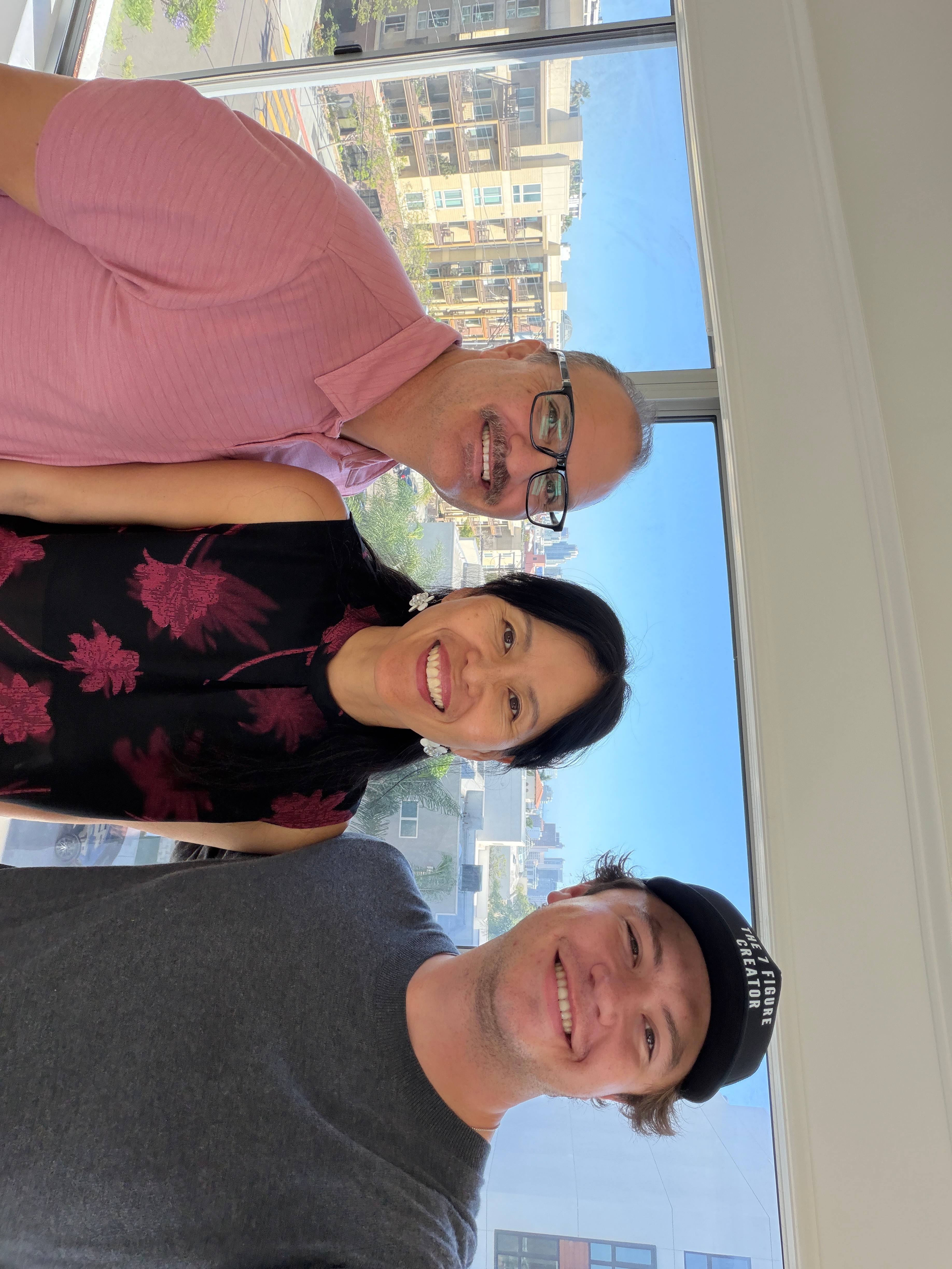 Three people smiling indoors with cityscape view through large windows behind them.