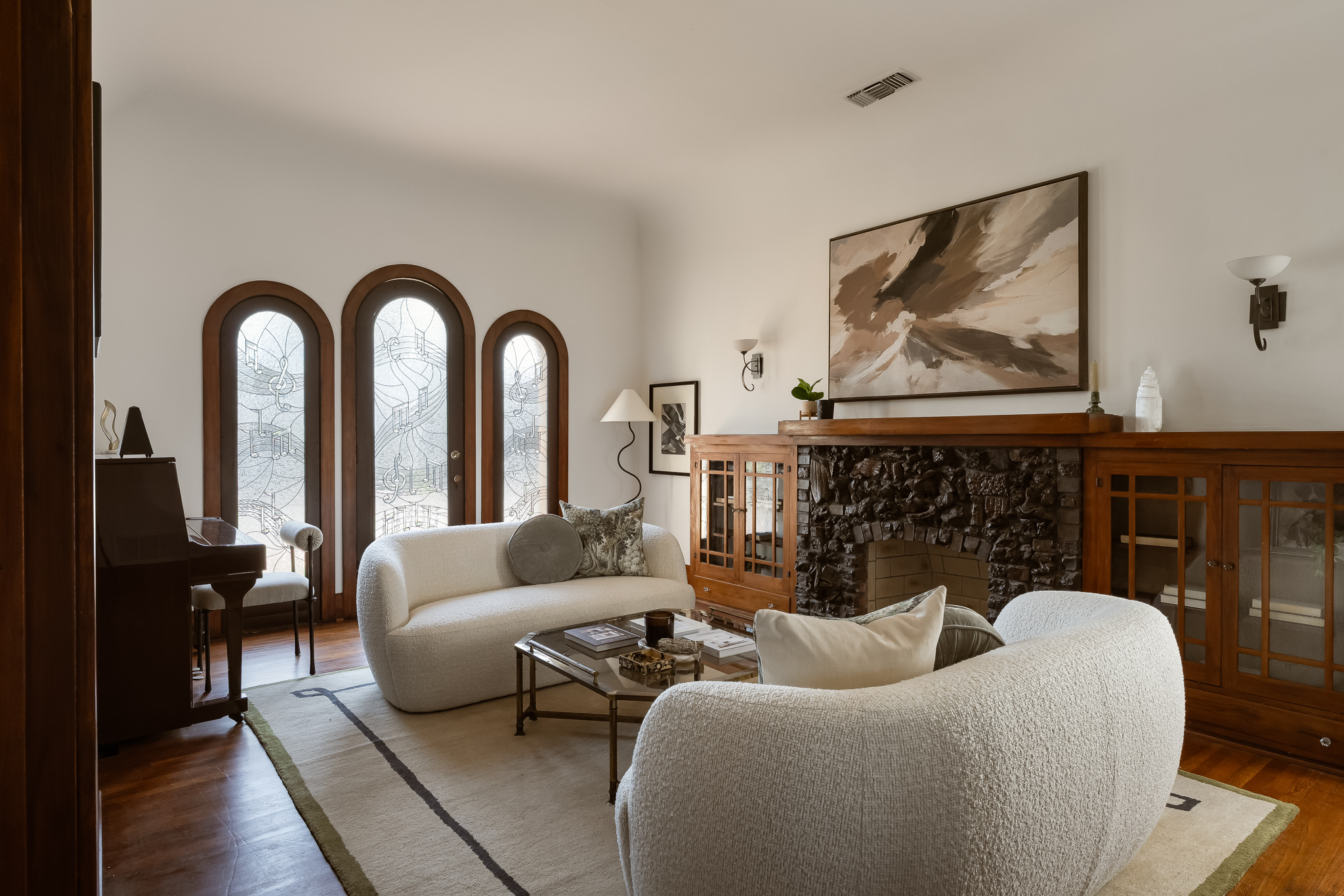 Living room with three arched windows, white sofa, armchair, wooden cabinet, and abstract painting, with a fireplace and wooden floor.