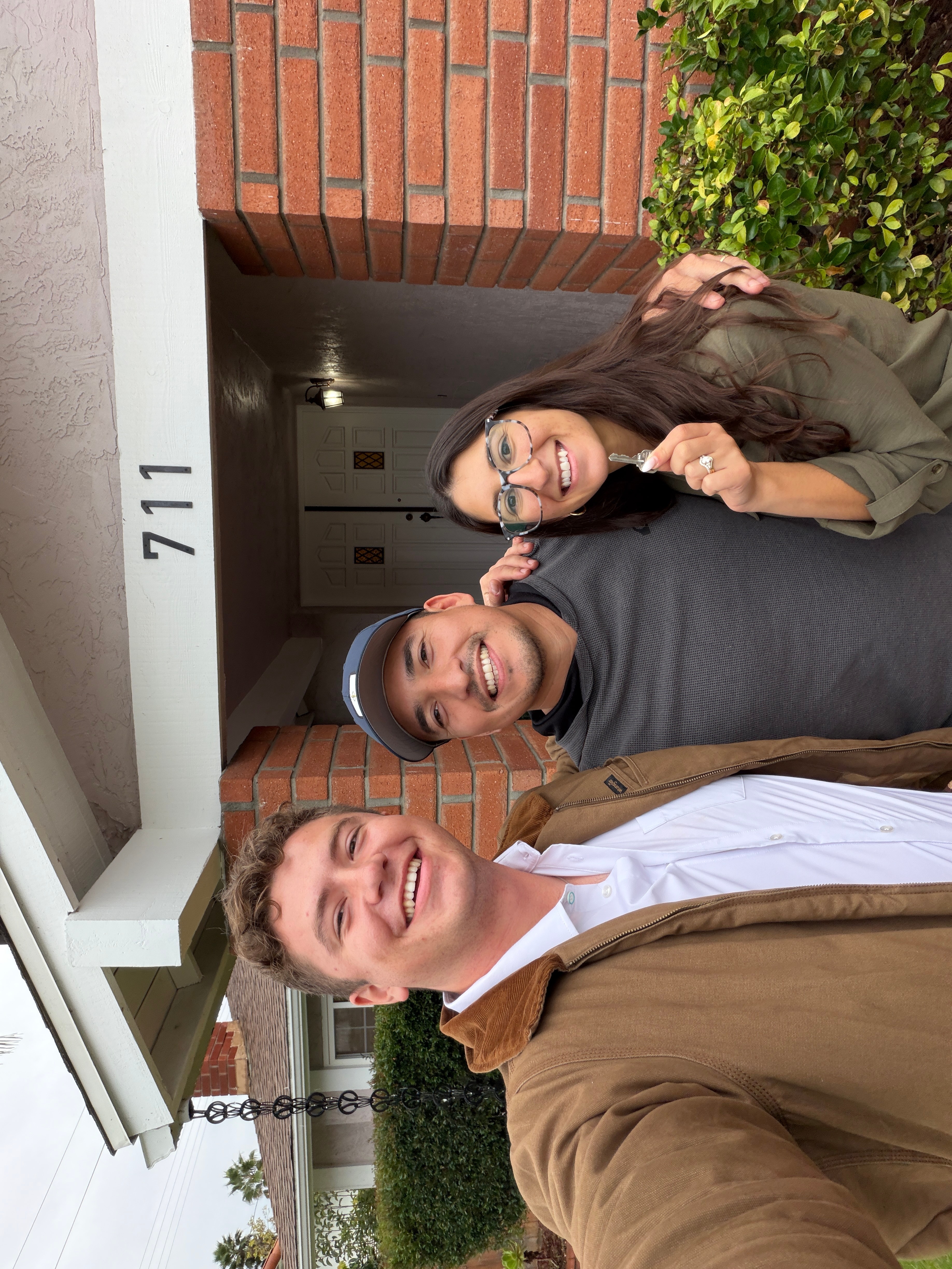 Three people smiling outside a building with the number 711, standing under a brick archway, with greenery in the background.