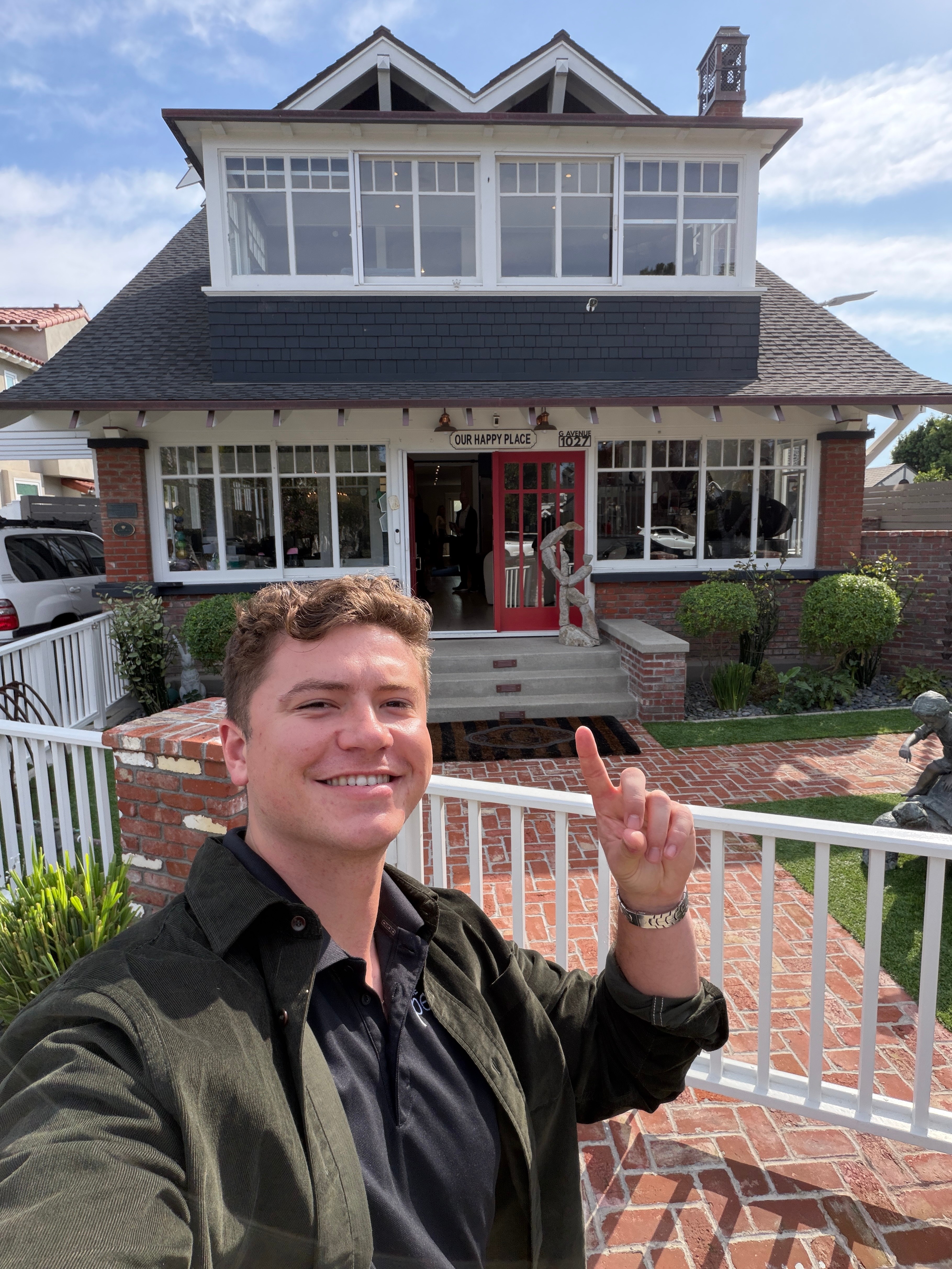 Young man smiling and pointing at a house with large windows and a porch, outdoors on a sunny day.