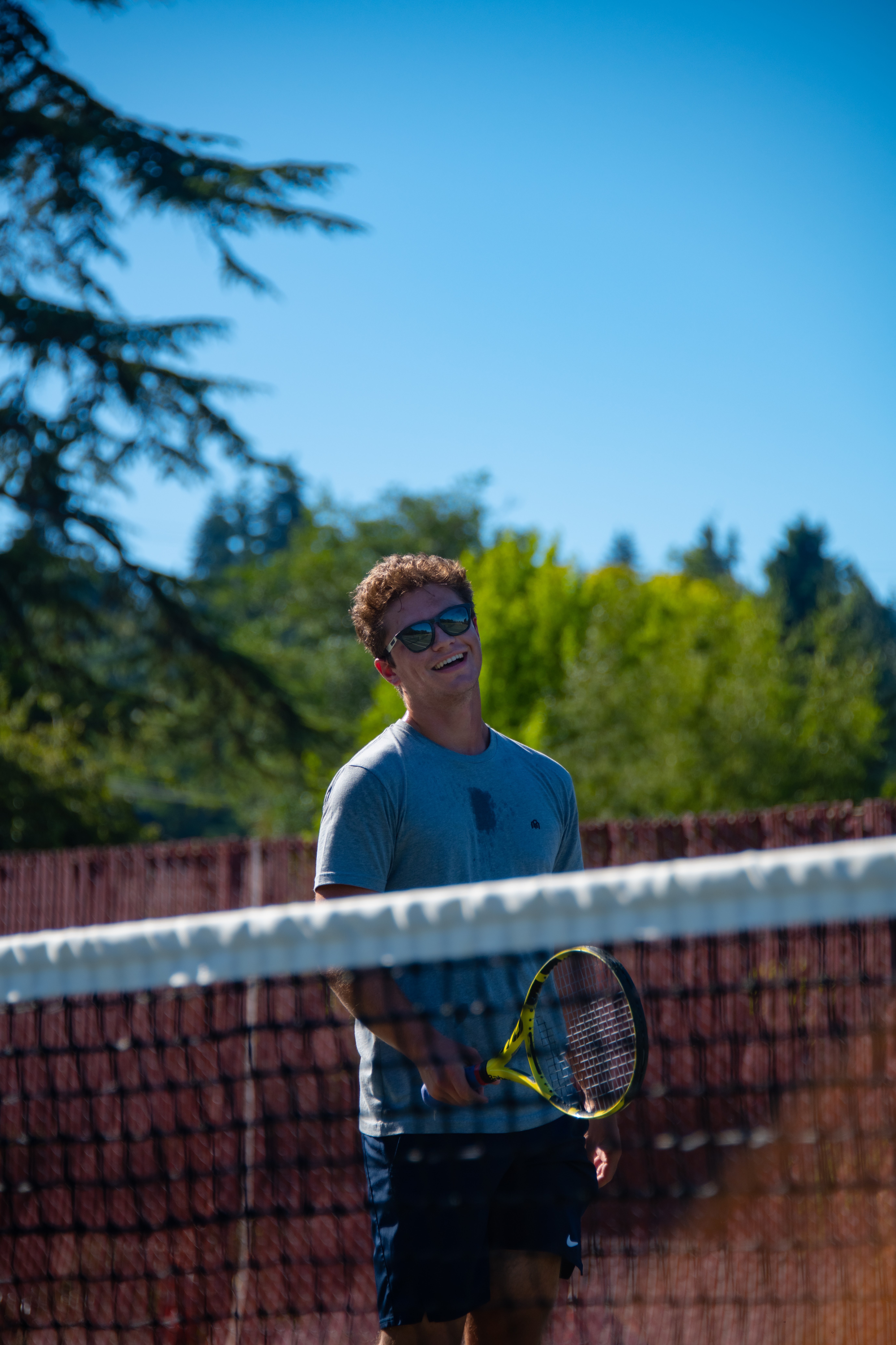 Young man holding a tennis racket on a court, wearing sunglasses and a blue shirt, with trees and blue sky in background.