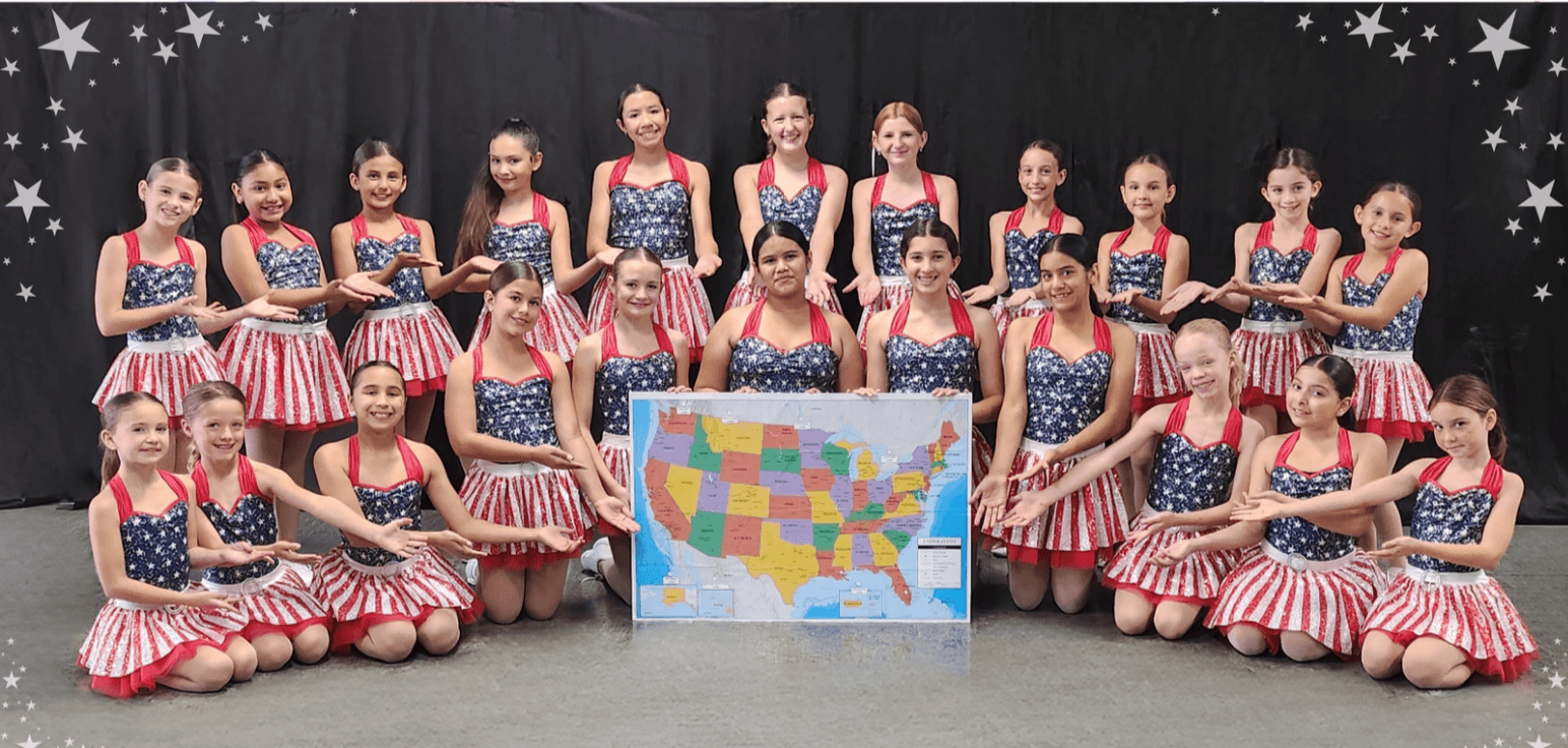 Group of young girls in patriotic costumes posing with a colorful map of the United States, standing and sitting in front of a black backdrop with stars.