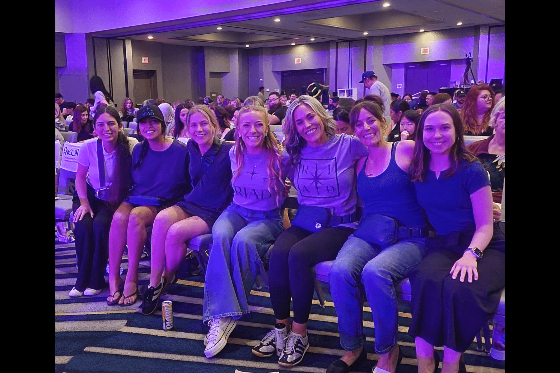 Group of eight women sitting together in a conference room, smiling, with purple lighting and audience in background.