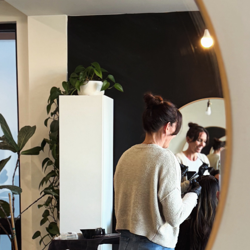 Two women in a salon, one styling hair, reflected in a mirror, with plants and a black wall in the background.