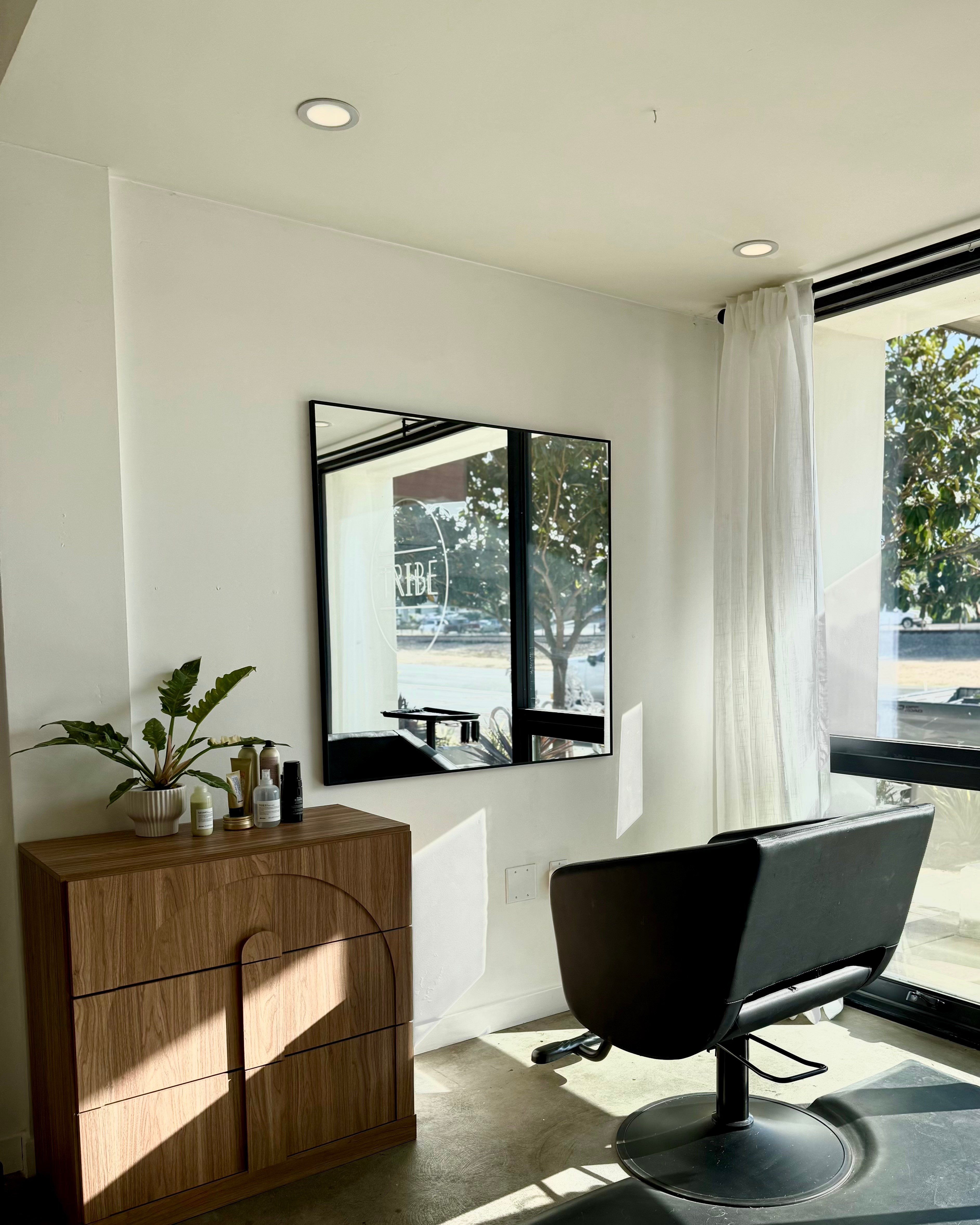 Interior of a room with a wooden cabinet, a mirror, a black chair, and a large window with white curtains, sunlight streaming in.