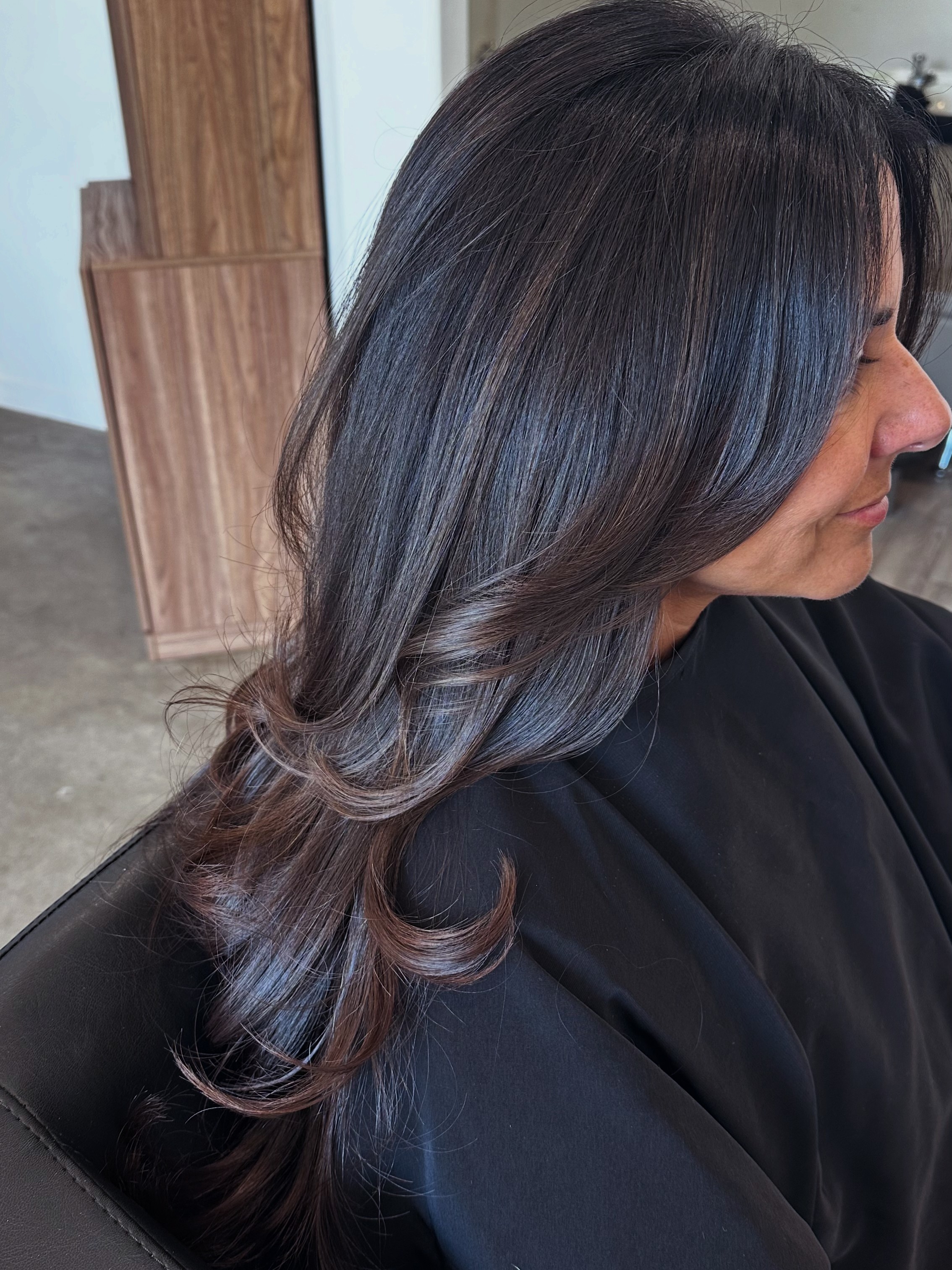 Side view of a woman with long, dark, wavy hair, sitting indoors with wooden furniture in the background.