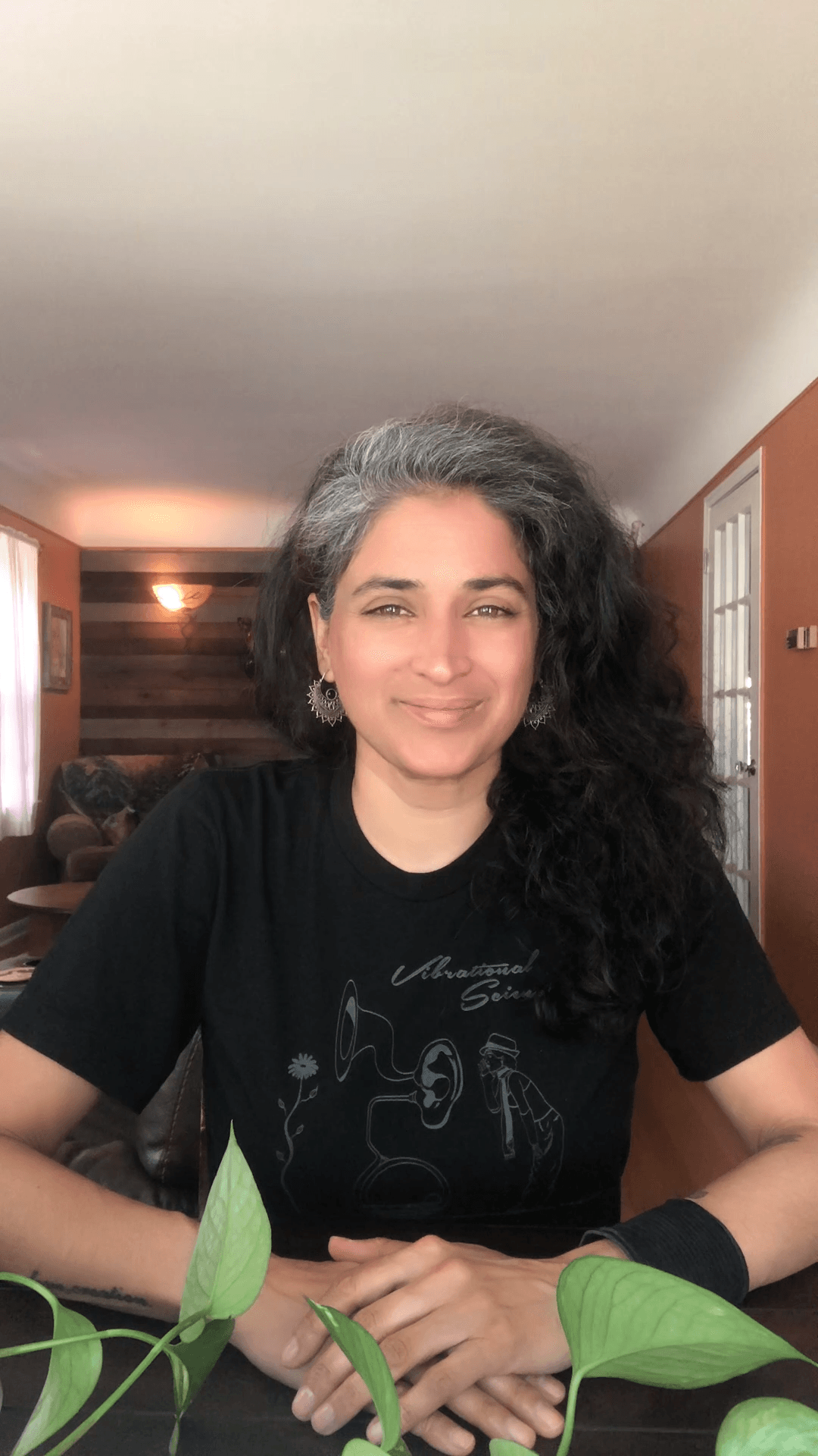 Woman with long, curly hair smiling, sitting at a table indoors, with a wooden wall and warm lighting in background.