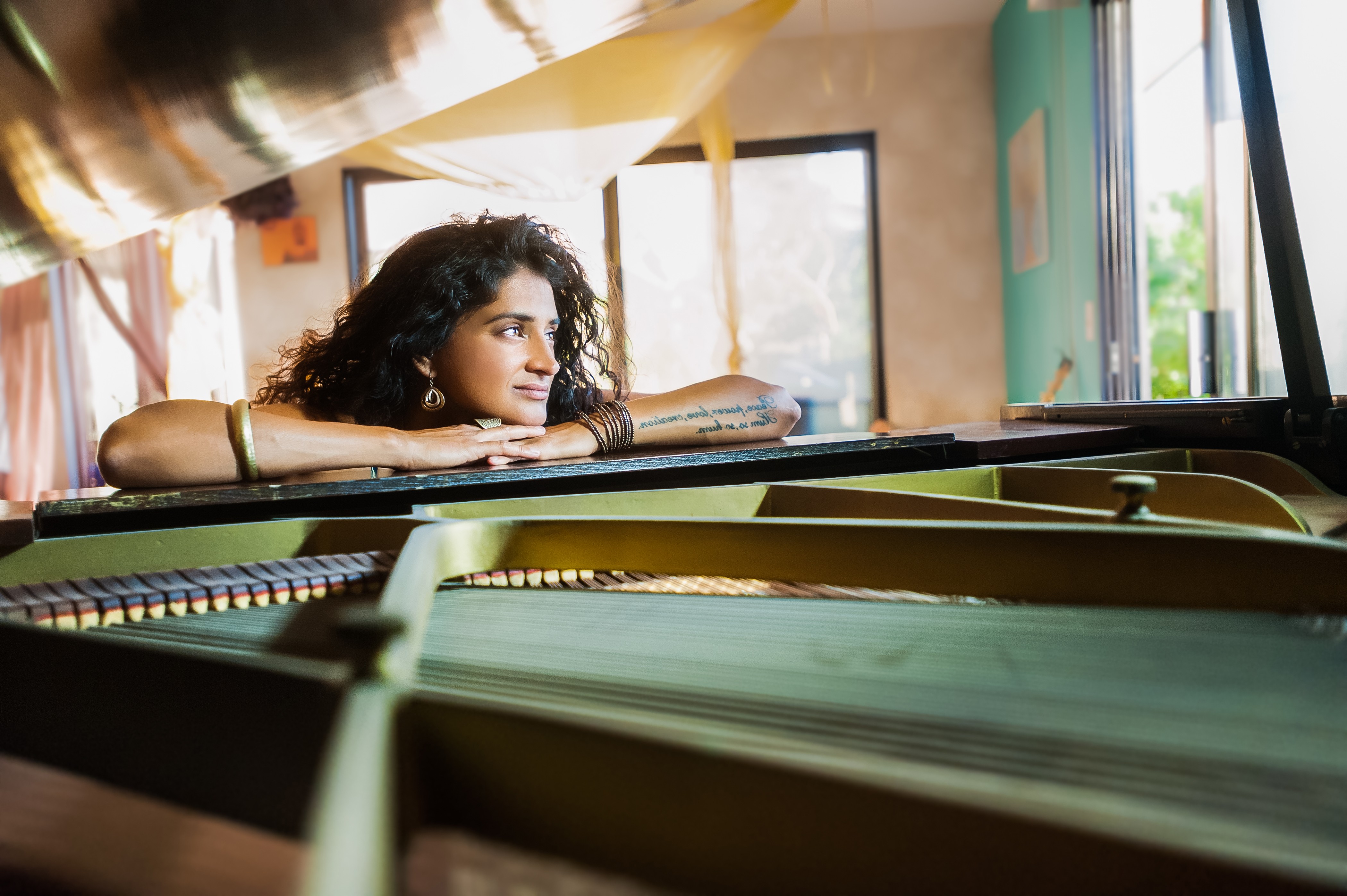 Young woman with curly hair resting her arms on a piano, looking out a window in a sunlit room.