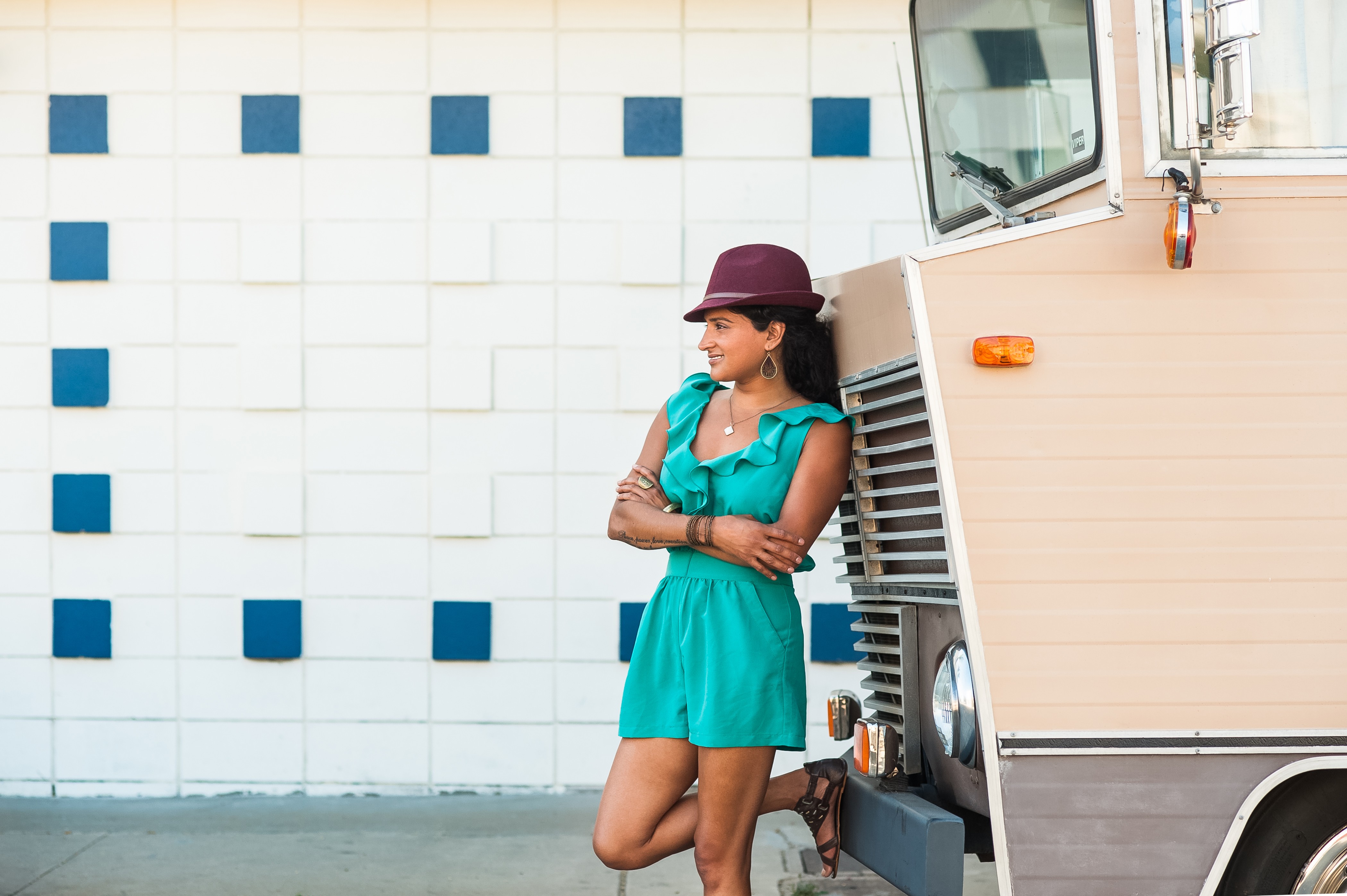 Woman in teal dress and hat leaning against a vehicle with crossed arms, standing outdoors near a tiled wall.