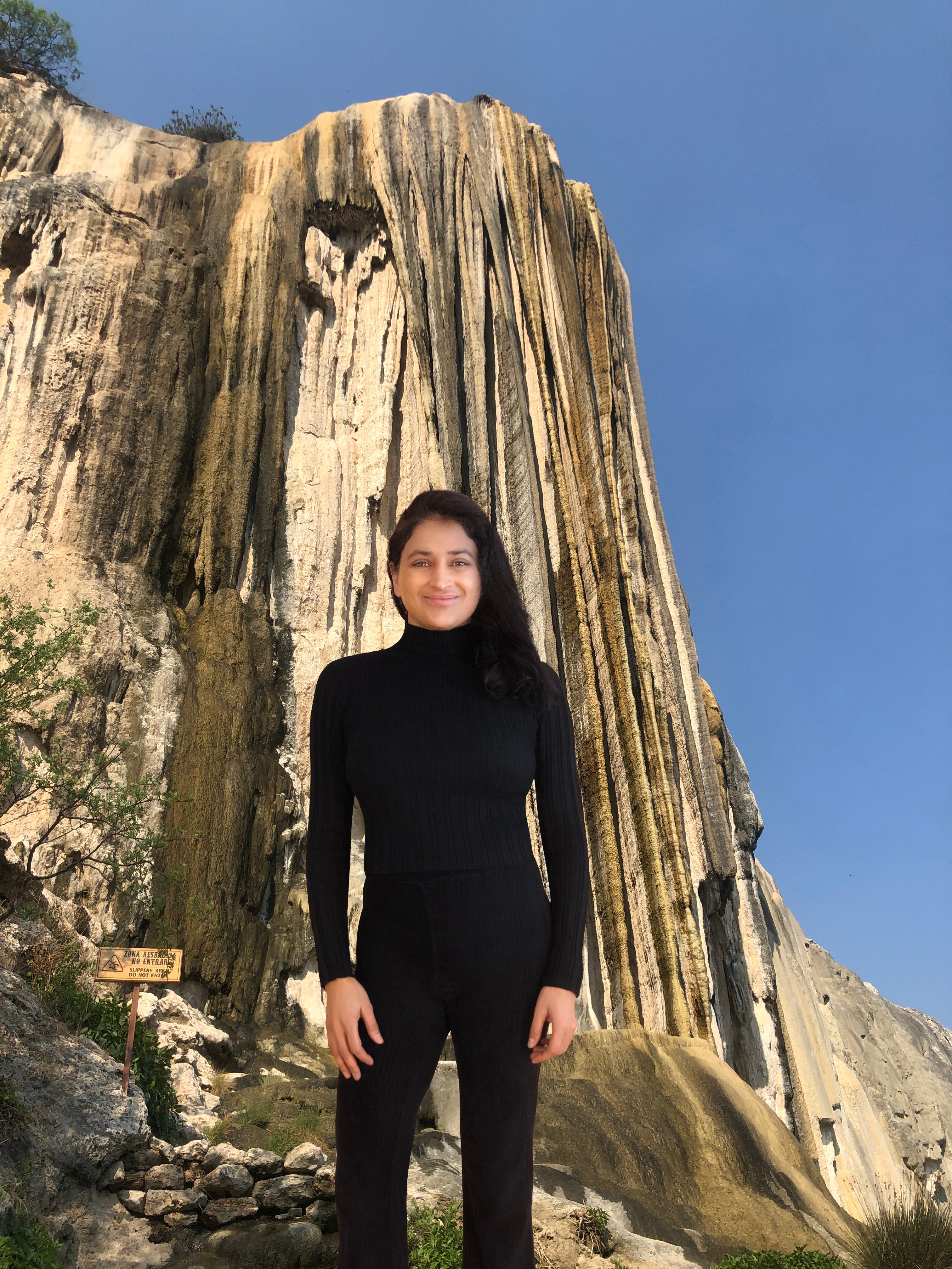 Young woman in black standing in front of a tall, textured rock formation under a clear blue sky.
