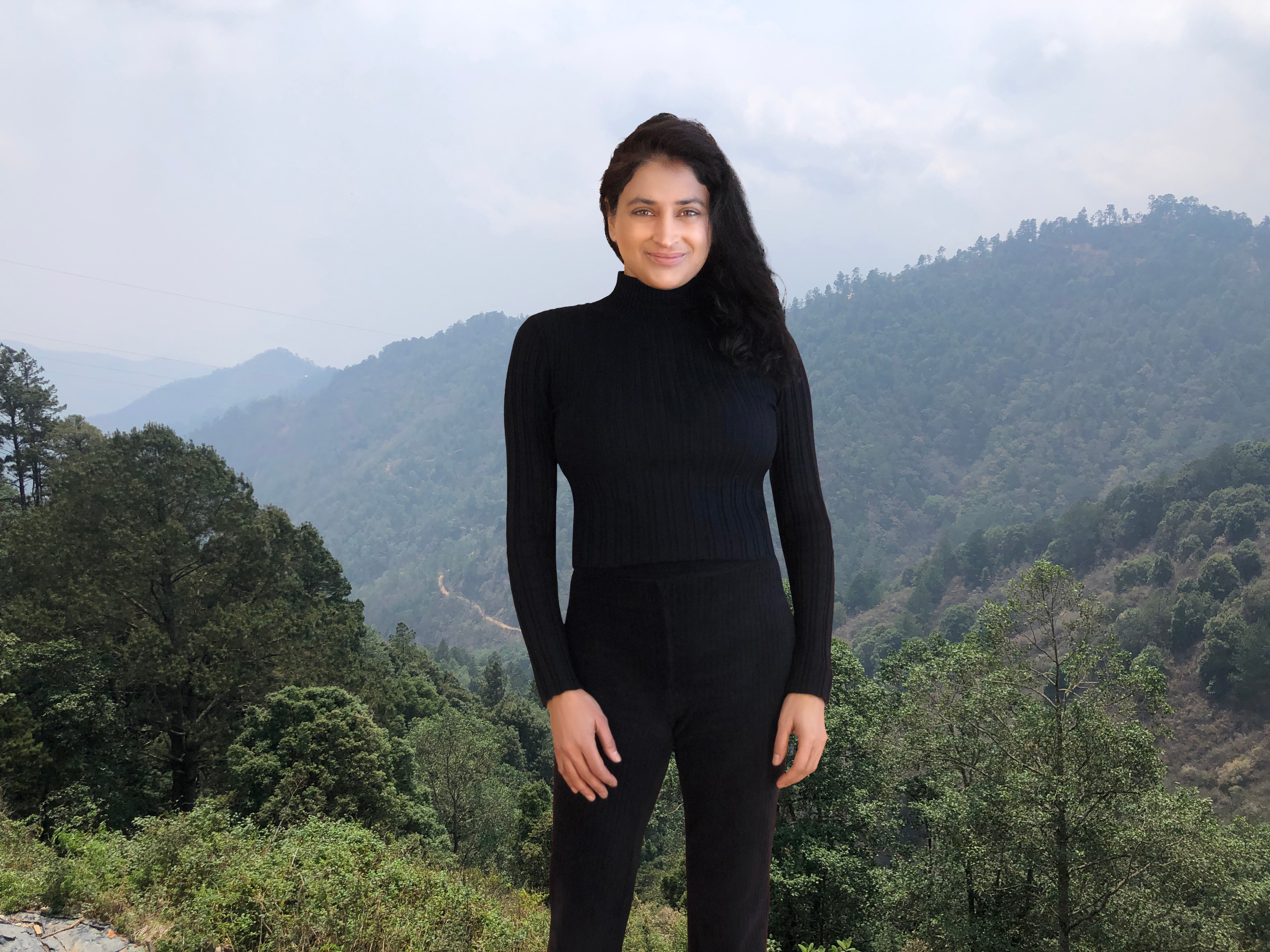 Woman in black standing outdoors with mountains and trees in the background.