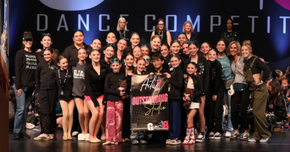Group of young dancers on stage holding a large banner, smiling, with a dark background and stage lights.