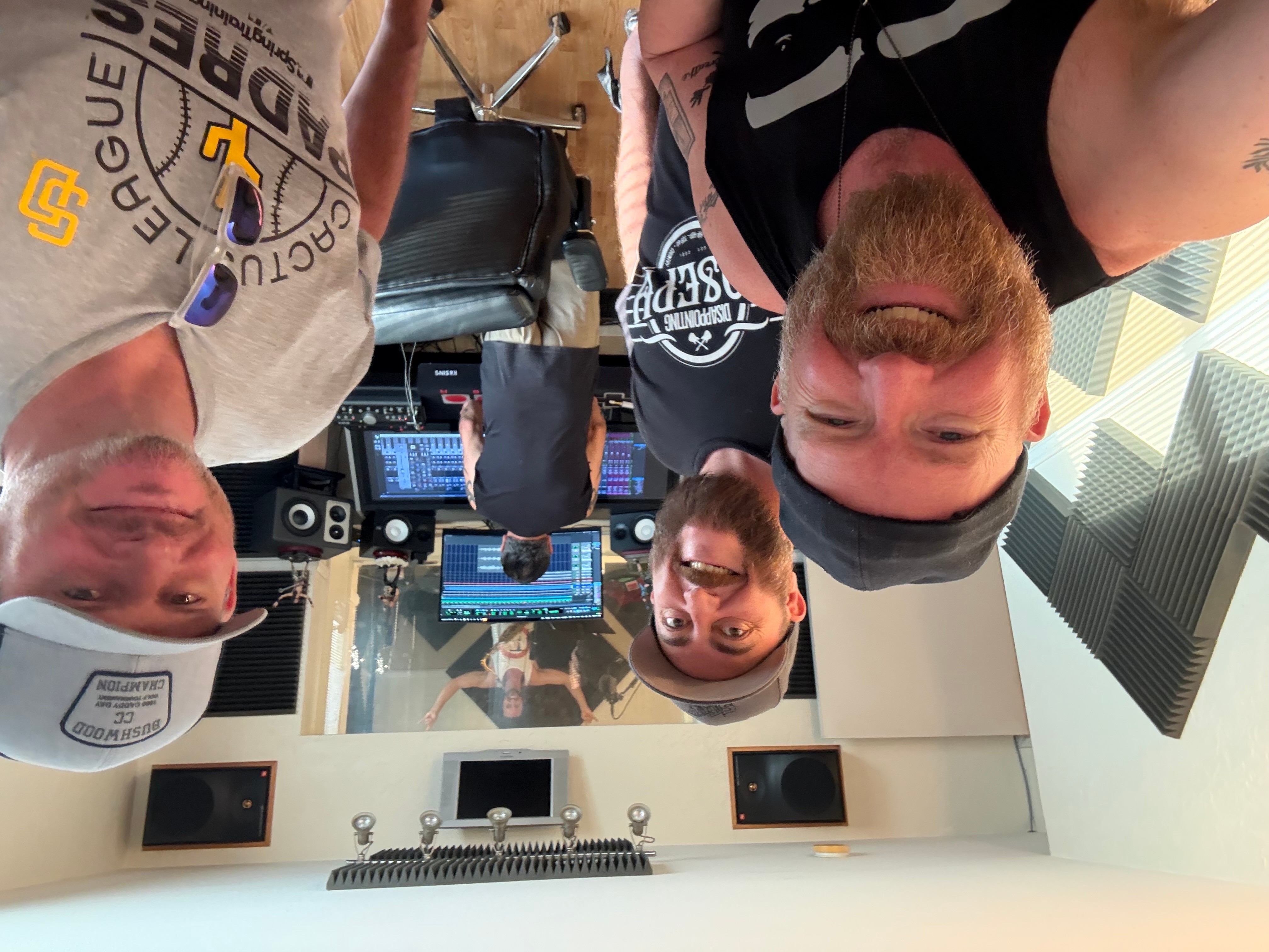 Four people in a recording studio, with equipment and soundproof walls, taking a selfie. One person wears a cap, others have glasses and beards.