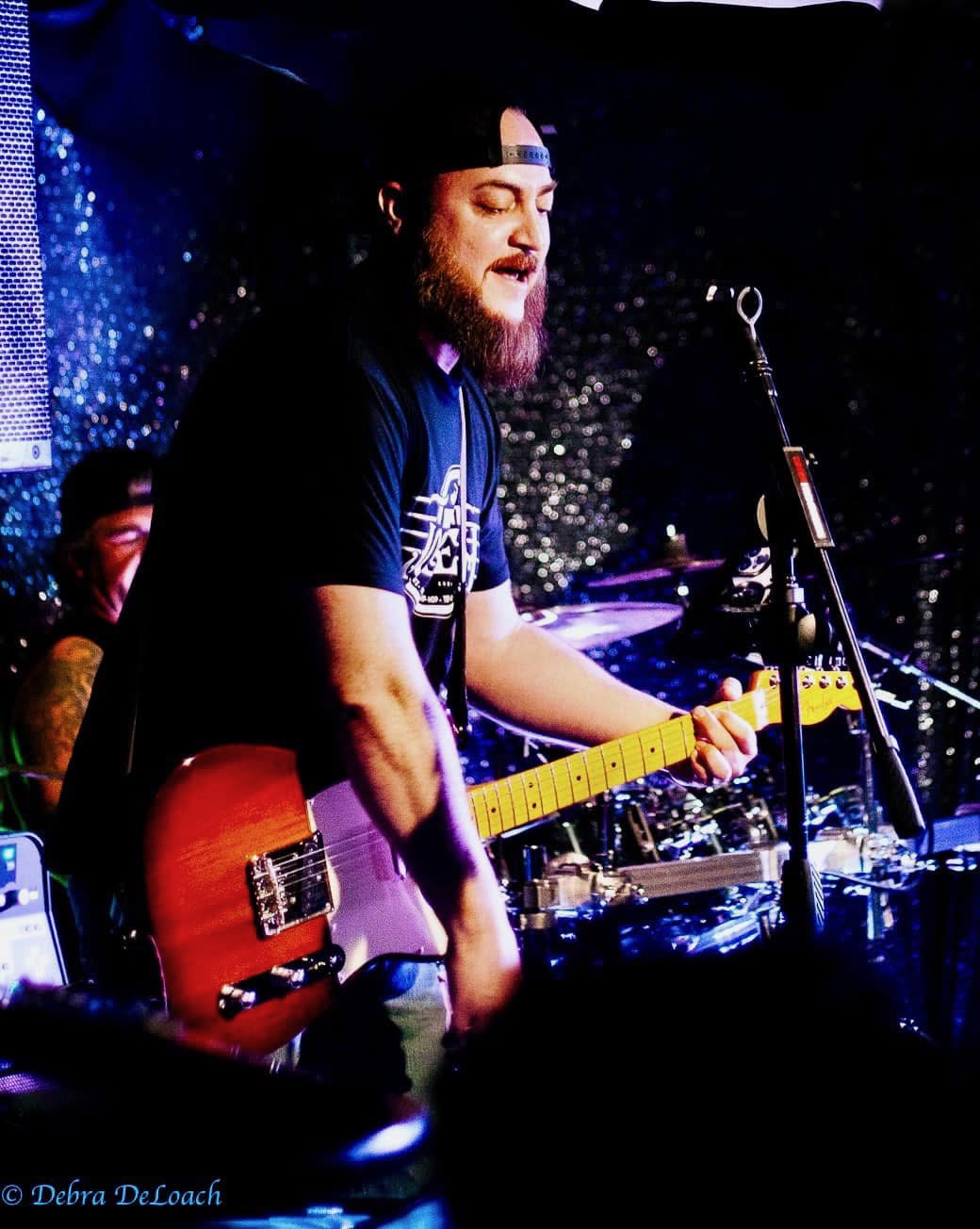 Person playing electric guitar on stage with drums and microphone, wearing a cap and dark clothing, performing live.