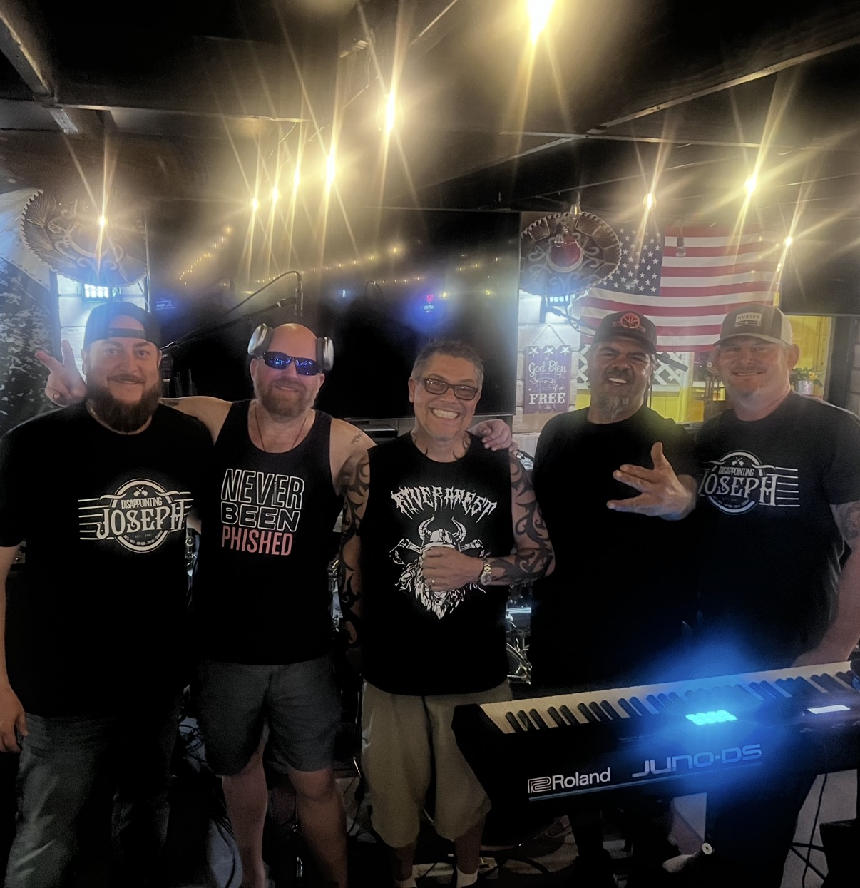 Group of five people standing behind a keyboard, smiling, in a dimly lit venue with bright lights and American flag in background.
