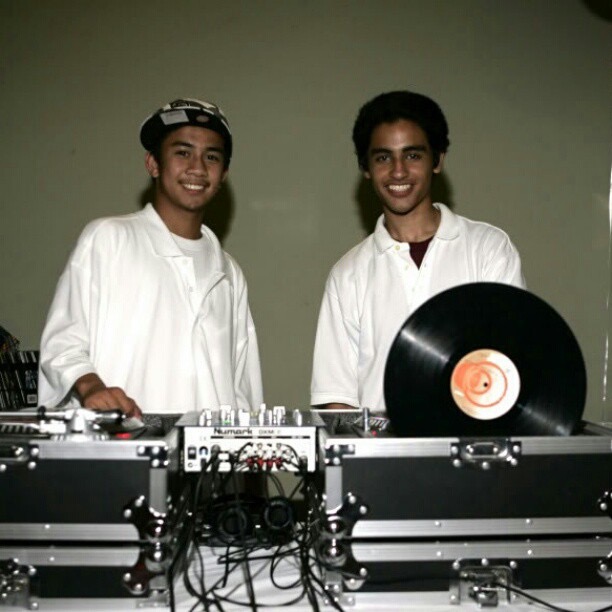 Two young people stand behind DJ equipment, smiling, with a vinyl record on a turntable, against a plain background.