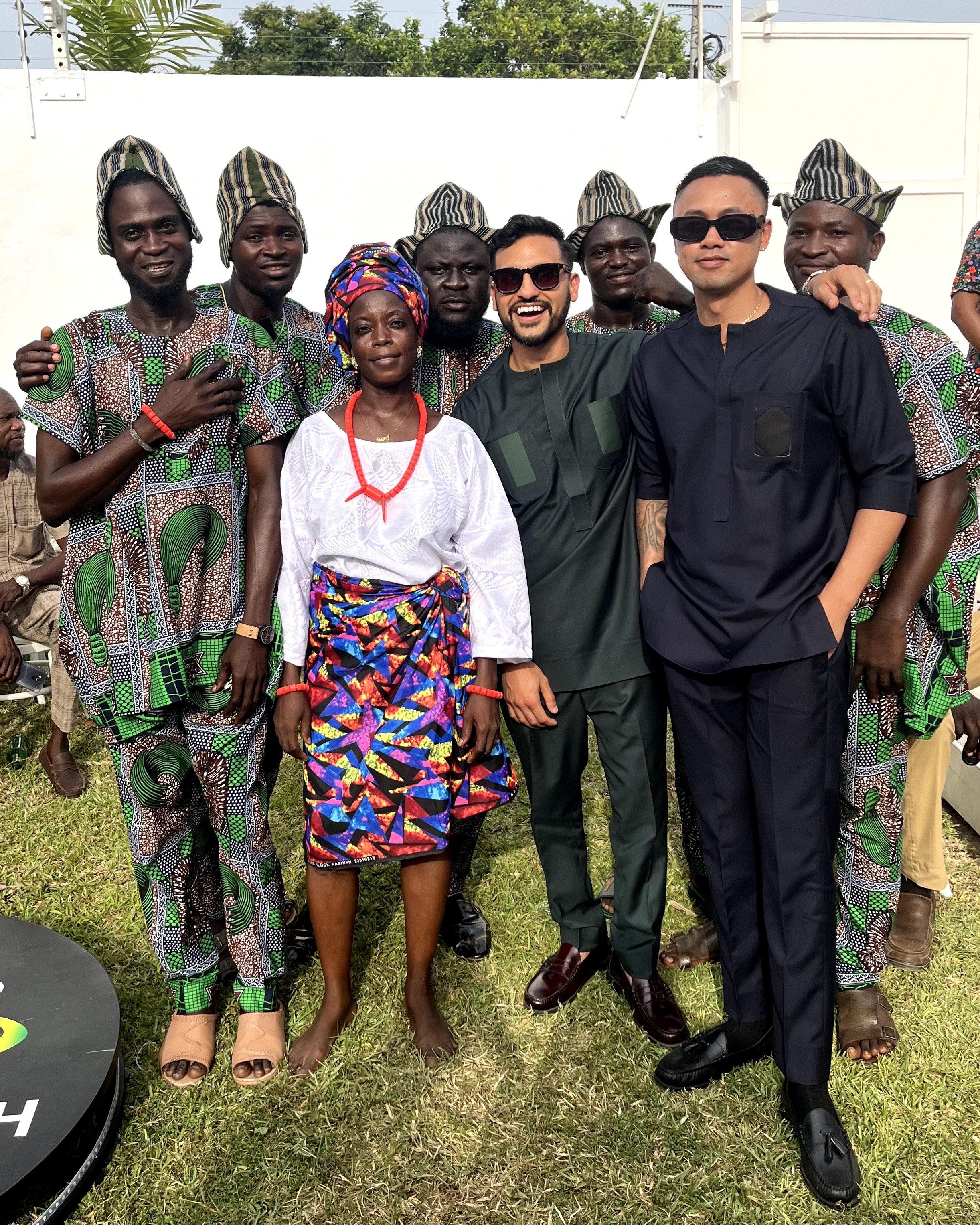 Group of nine people outdoors, some wearing traditional African clothing and others in modern attire, standing on grass.