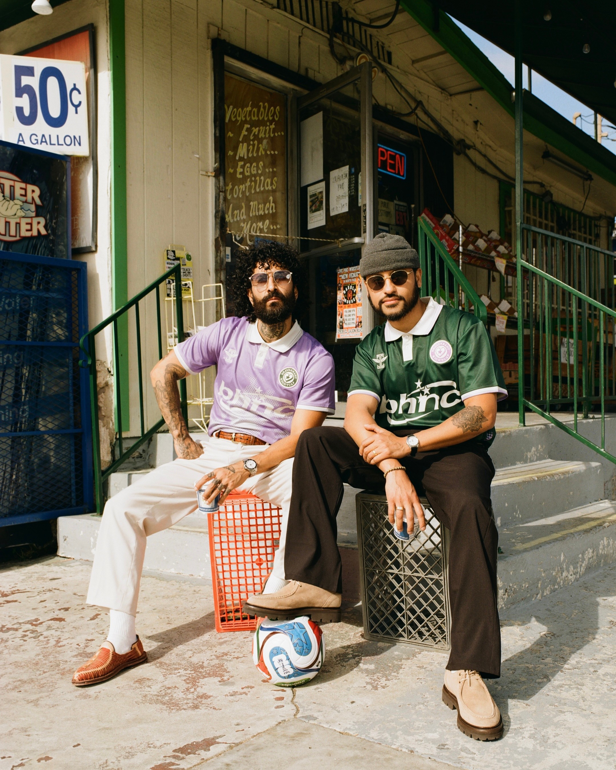 Two men sit on steps outside a small shop, one with a beard and glasses, the other wearing a cap, with a soccer ball between them.