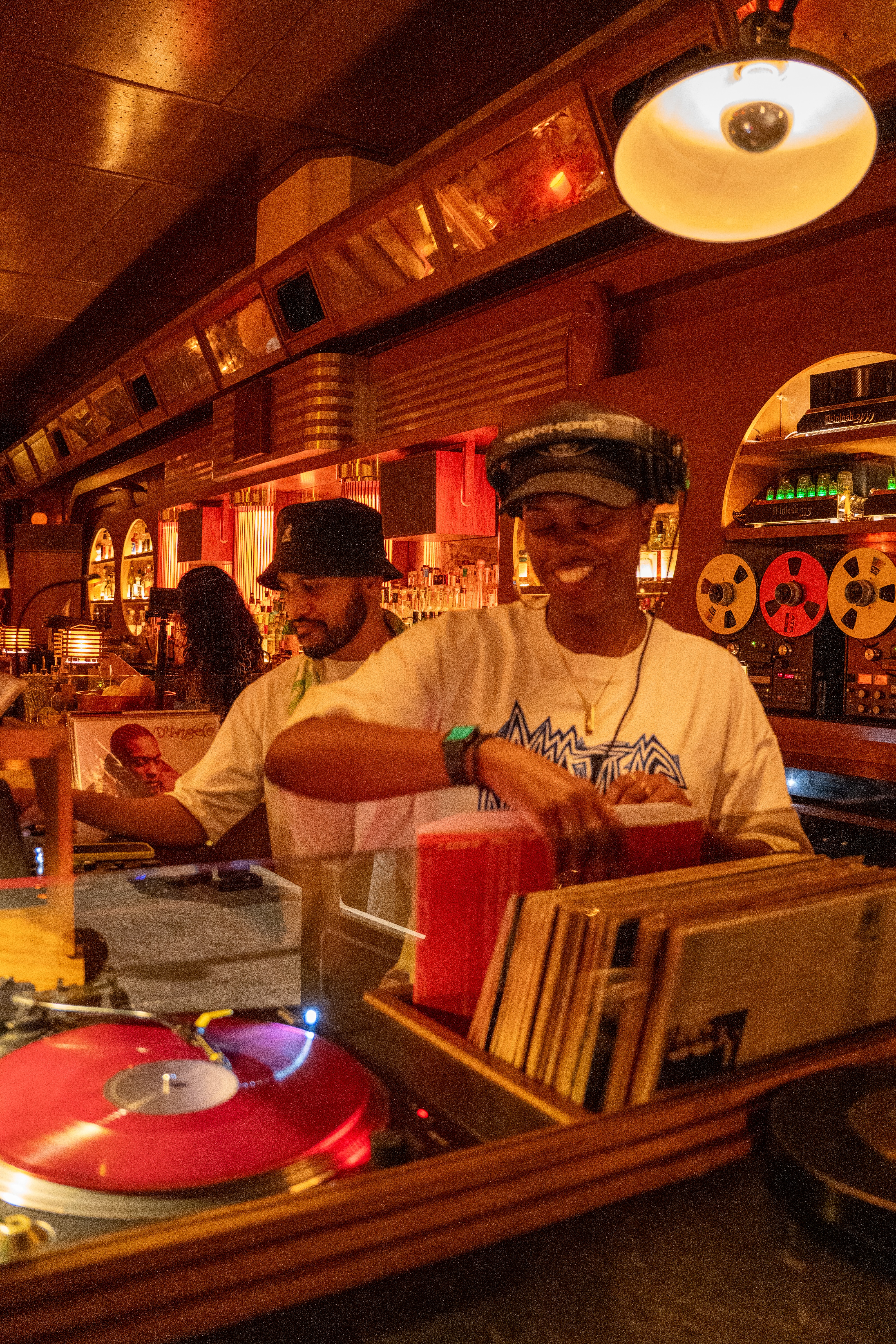 Two people behind a bar counter, smiling, with shelves of bottles and glasses in the background.