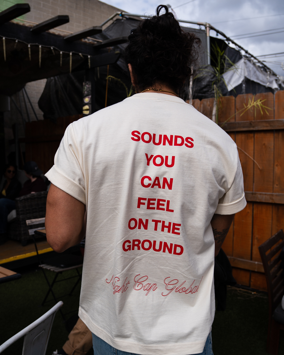 Person with curly hair wearing a white T-shirt with red text on the back, outdoors with a wooden fence and canopy.