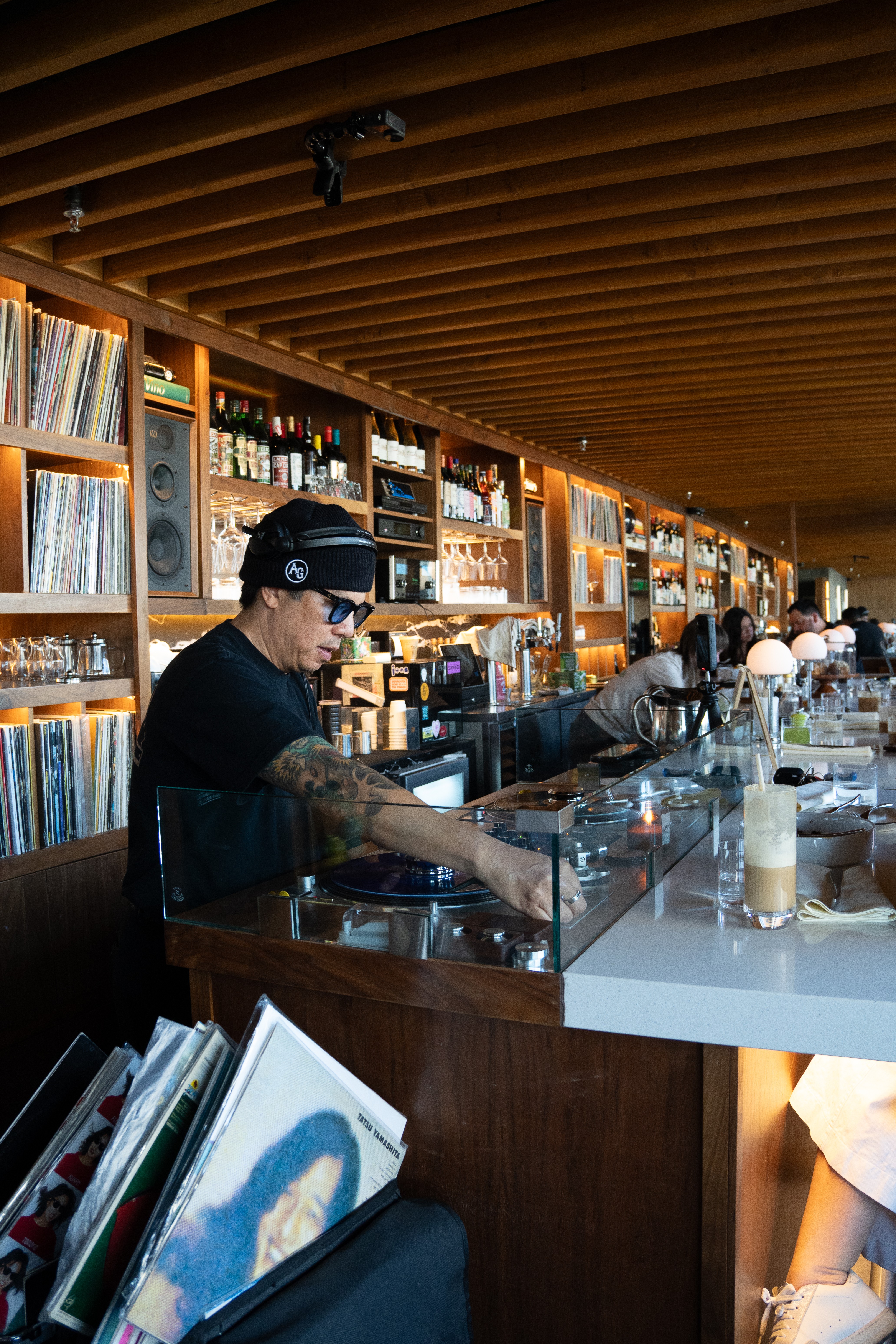 Barista preparing drinks at a modern bar with shelves of books and bottles, warm lighting, and a wooden ceiling.