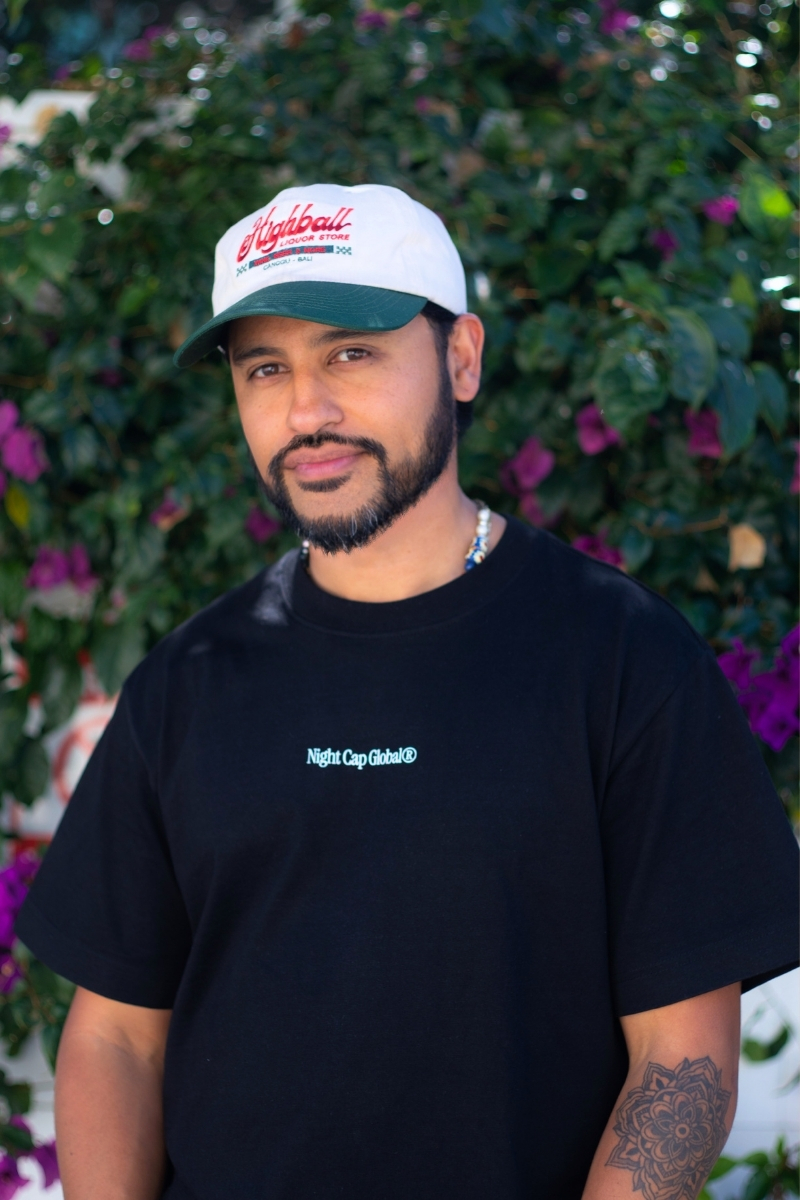 Young man with beard and mustache wearing a white cap and black T-shirt, standing outdoors with flowering bushes in background.