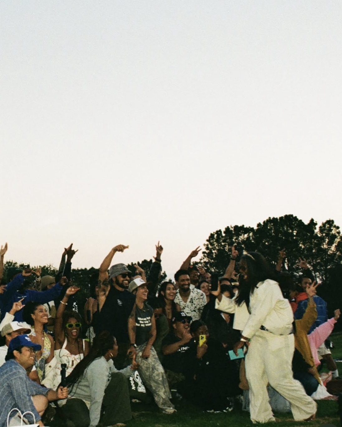 Group of people outdoors, some raising hands, others taking photos, with trees and sky in background.