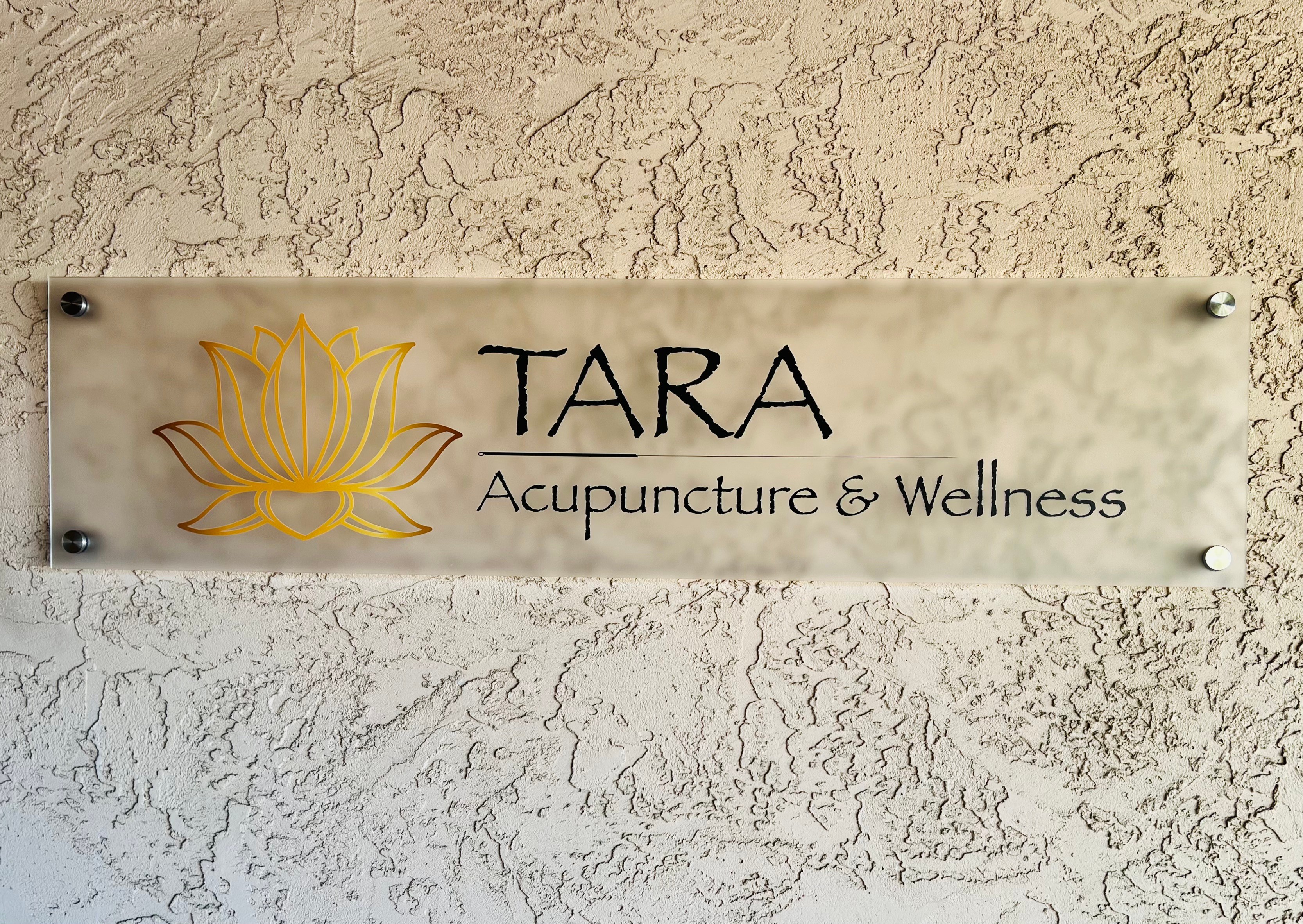 Sign with a yellow lotus flower, text reads 'TARA Acupuncture & Wellness', mounted on a textured wall.