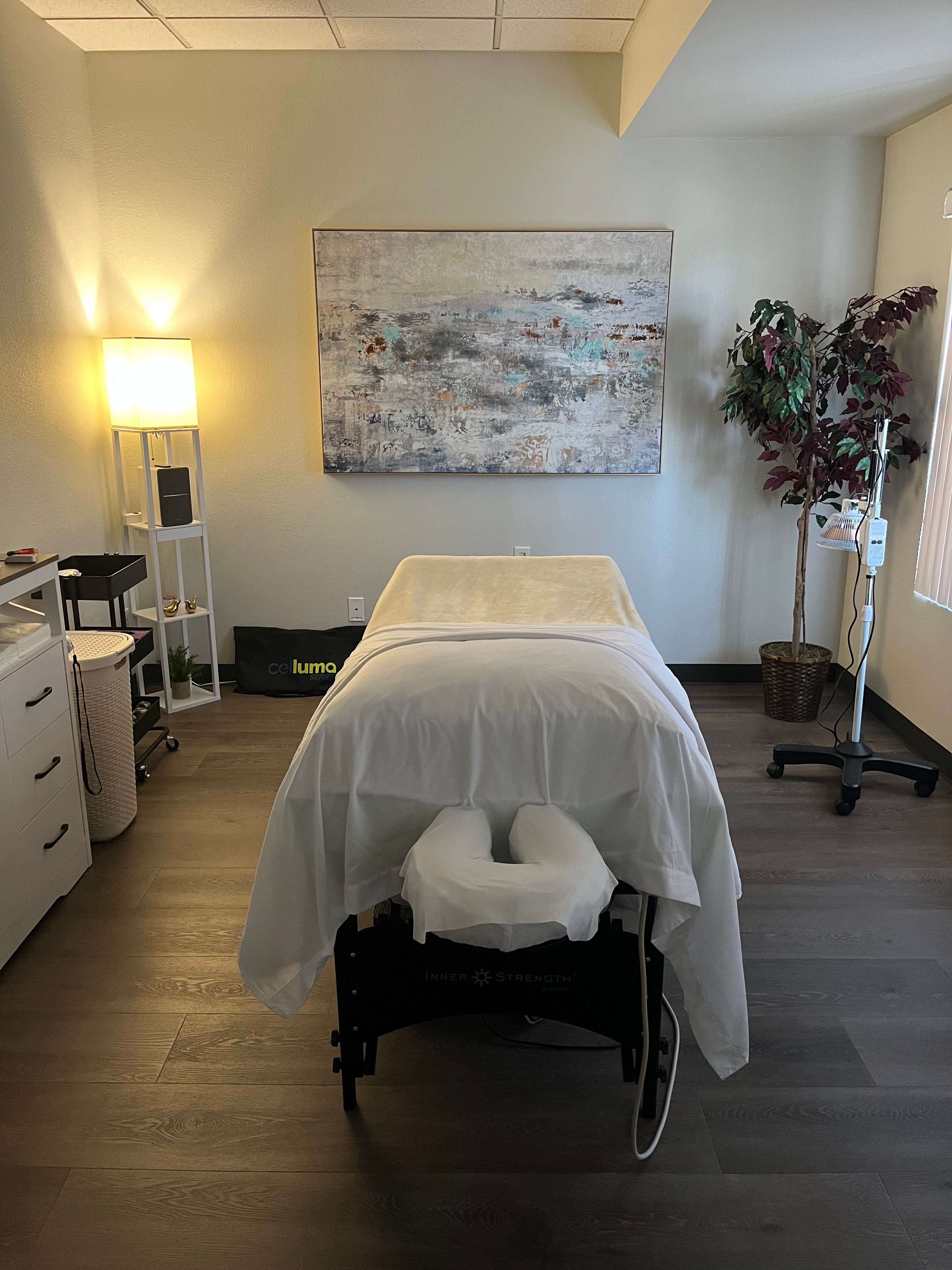 Treatment room with a massage table, abstract wall art, a lamp, and a potted plant, with natural light from a window.