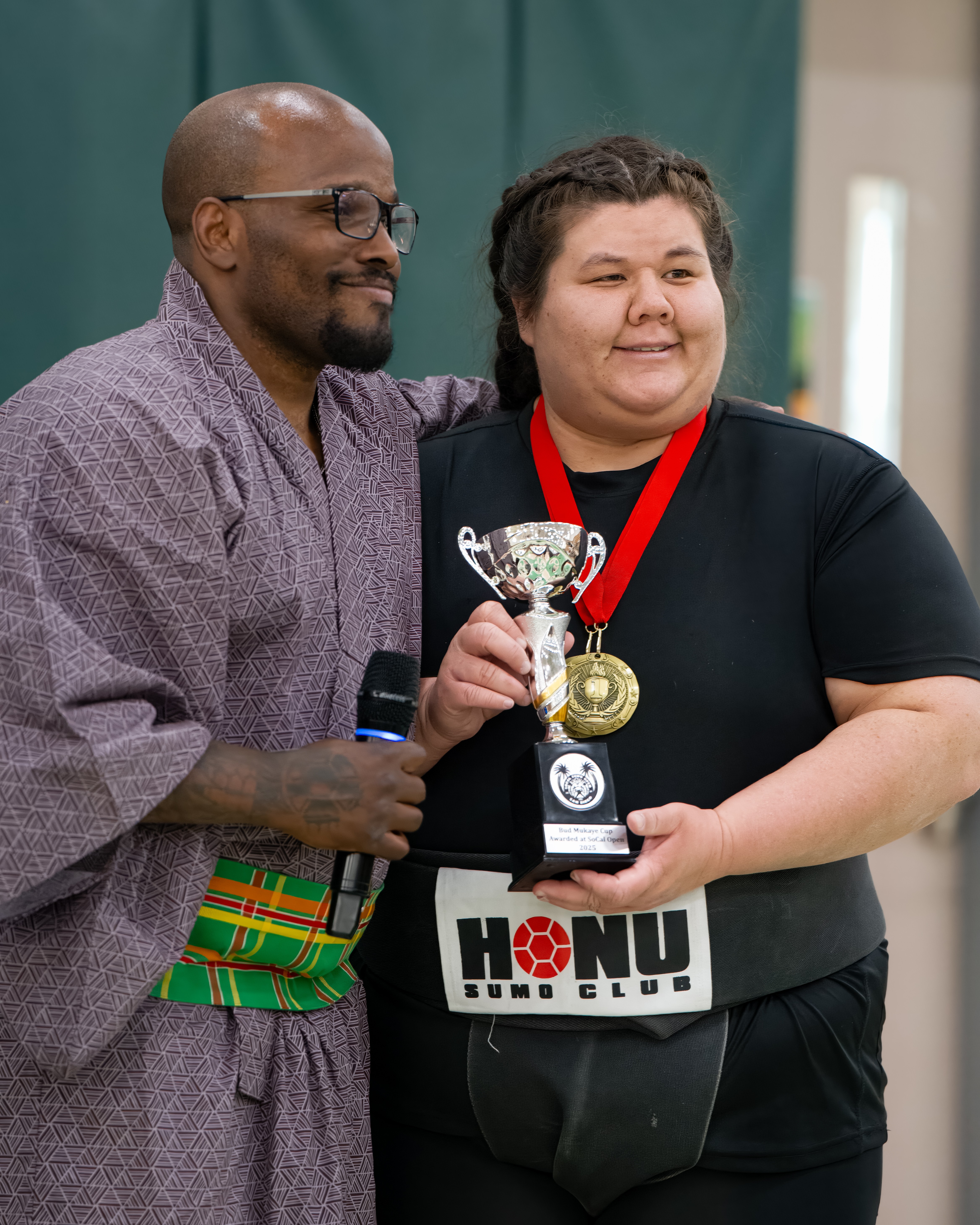 Two people smiling, one holding a trophy and wearing medals, in an indoor setting.