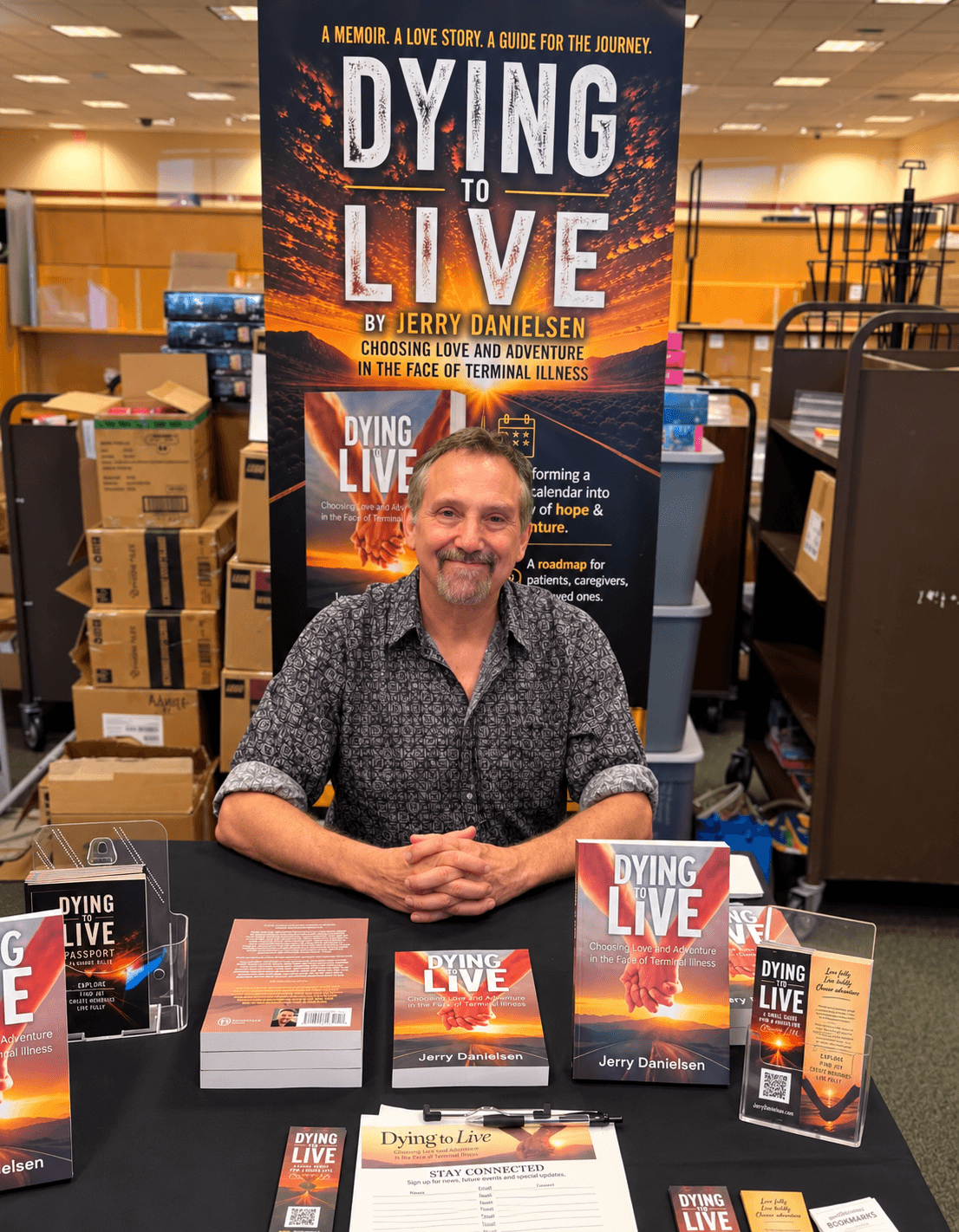 Man sitting at a table with books and promotional materials for 'Dying to Live' in a bookstore, shelves in background.