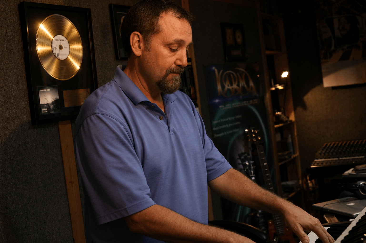 Man in blue shirt playing keyboard in recording studio with gold record on wall.