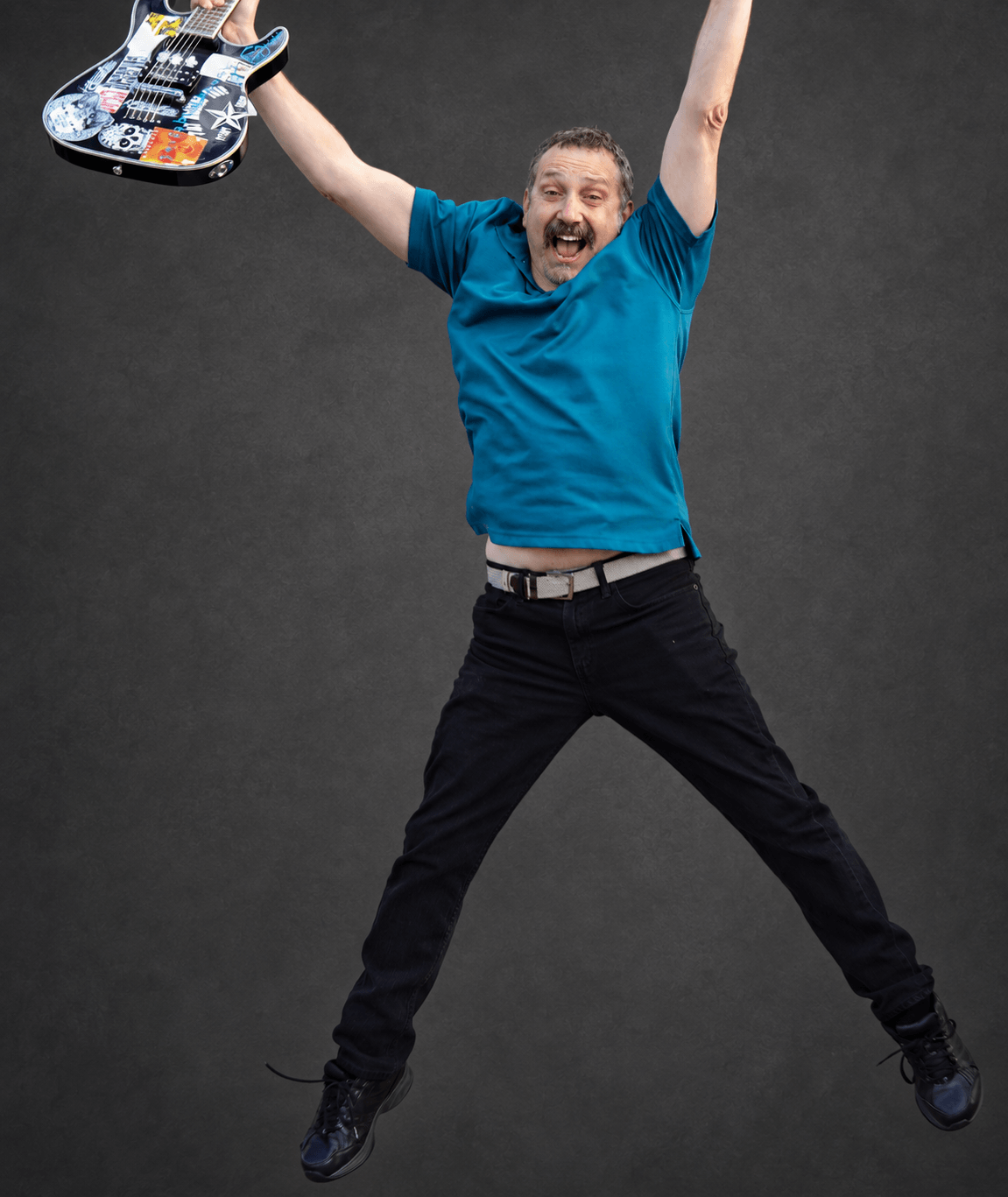 Man jumping with arms raised, holding a decorated guitar, smiling, against a dark background.