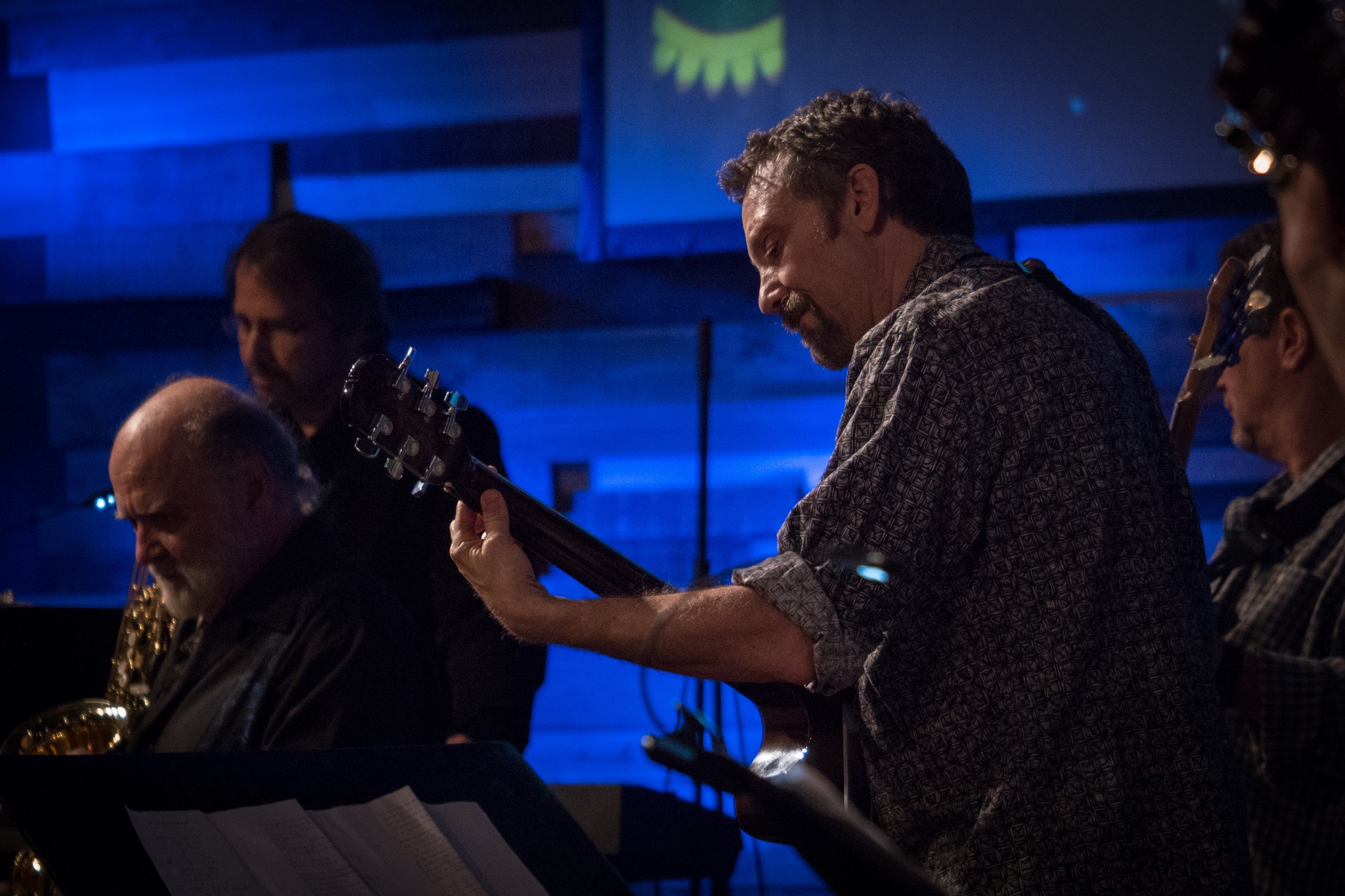 Musicians perform on stage with blue lighting, including a man playing guitar and others in the background.
