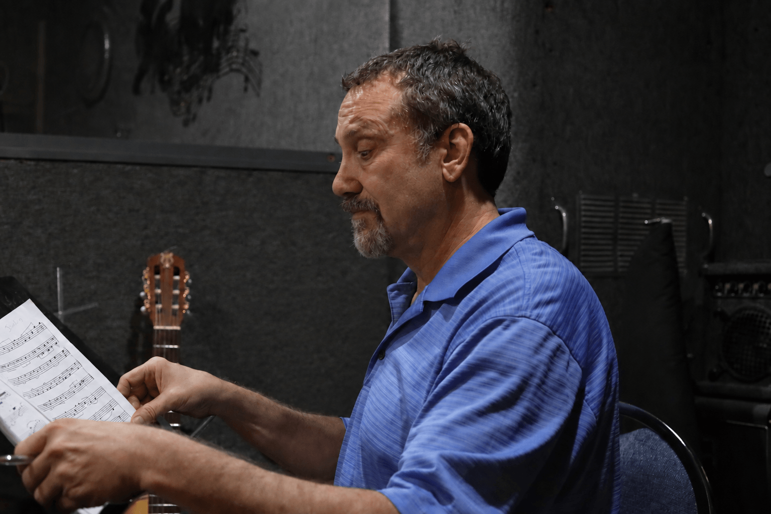 Man with gray hair and beard reading sheet music in a dark room, wearing a blue shirt.