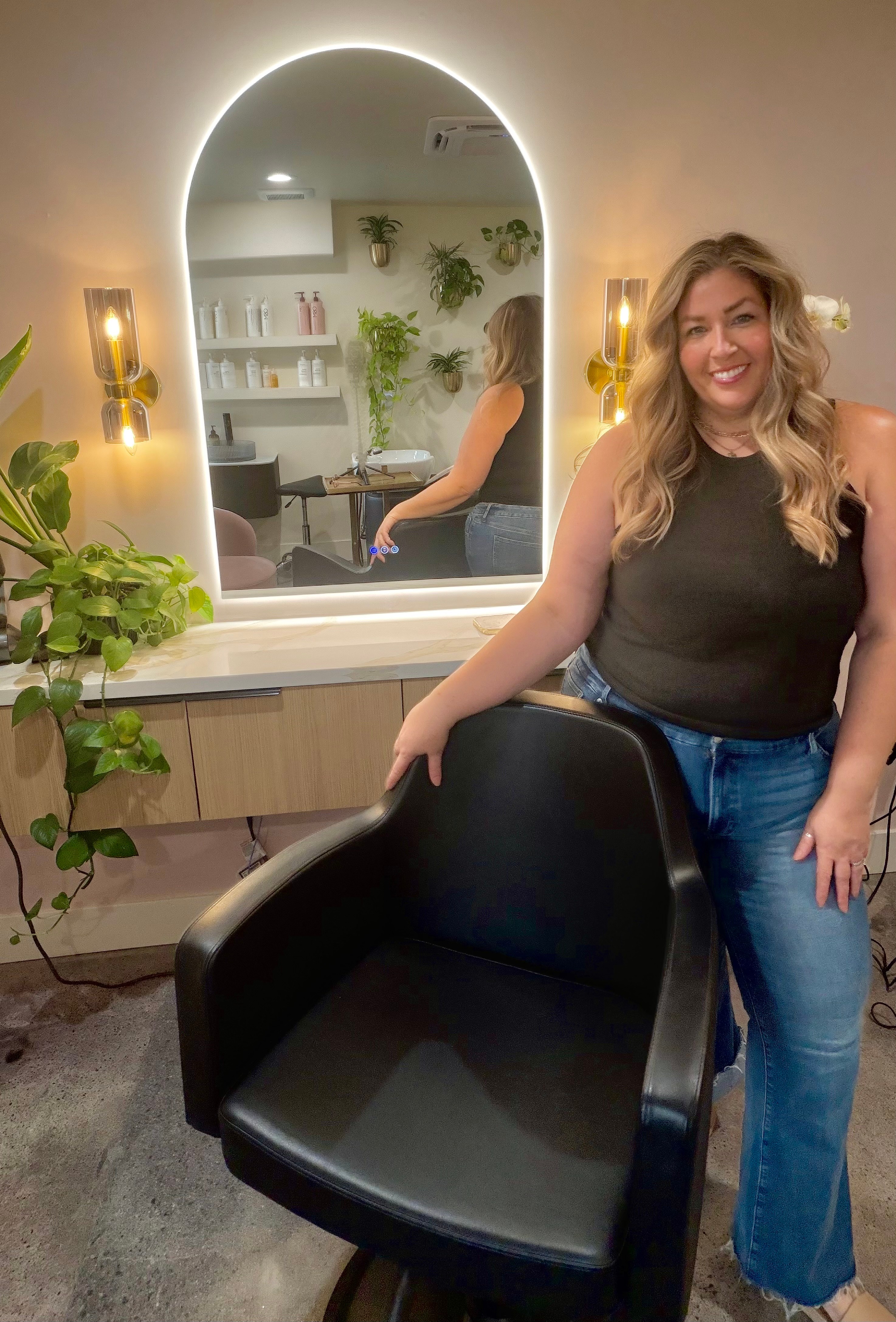 Woman with long blonde hair smiling, standing next to a black salon chair in a hair salon, with a mirror and plants in the background.