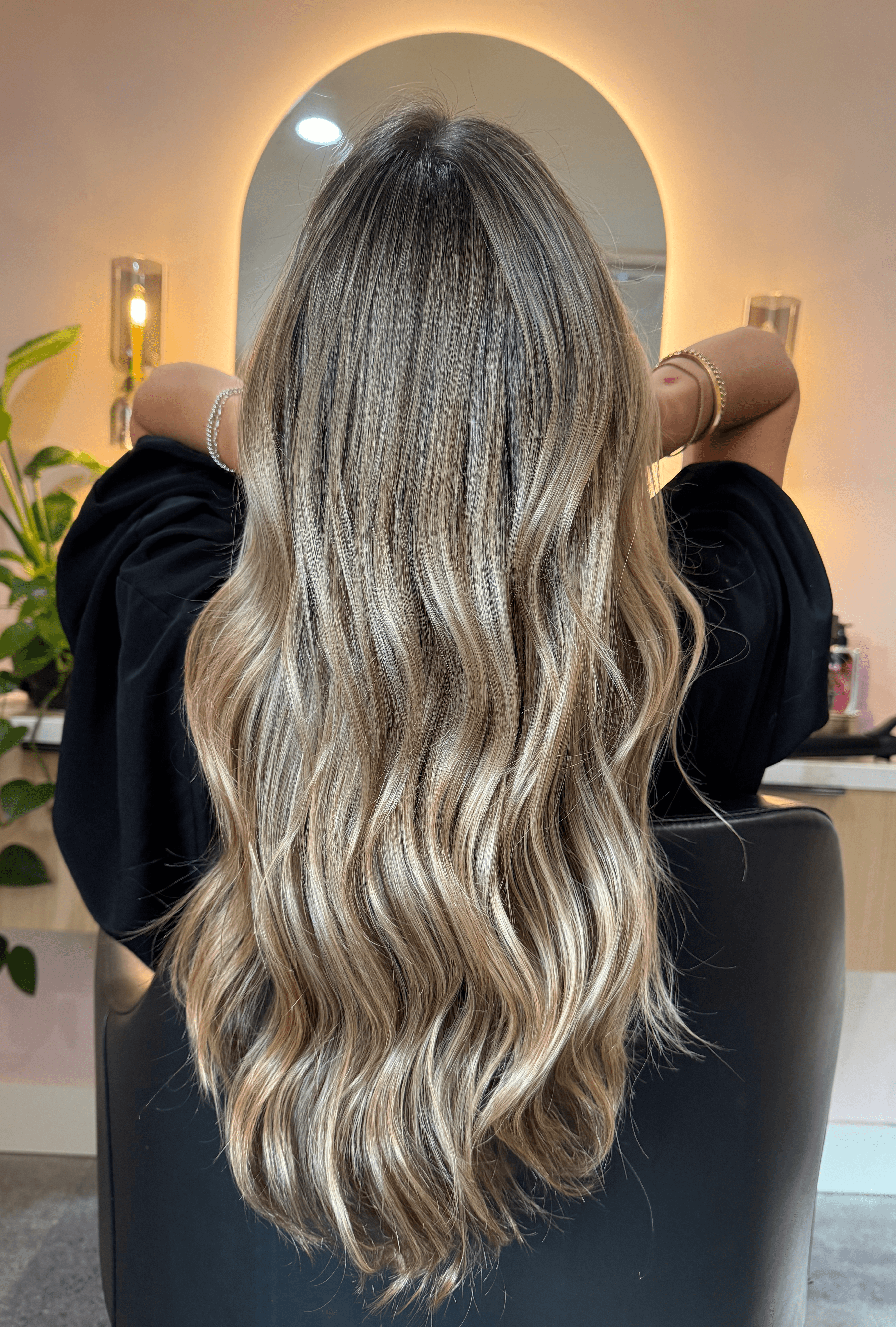 Person with long, wavy blonde hair facing mirror in a salon, arms raised, with plants and salon tools visible.