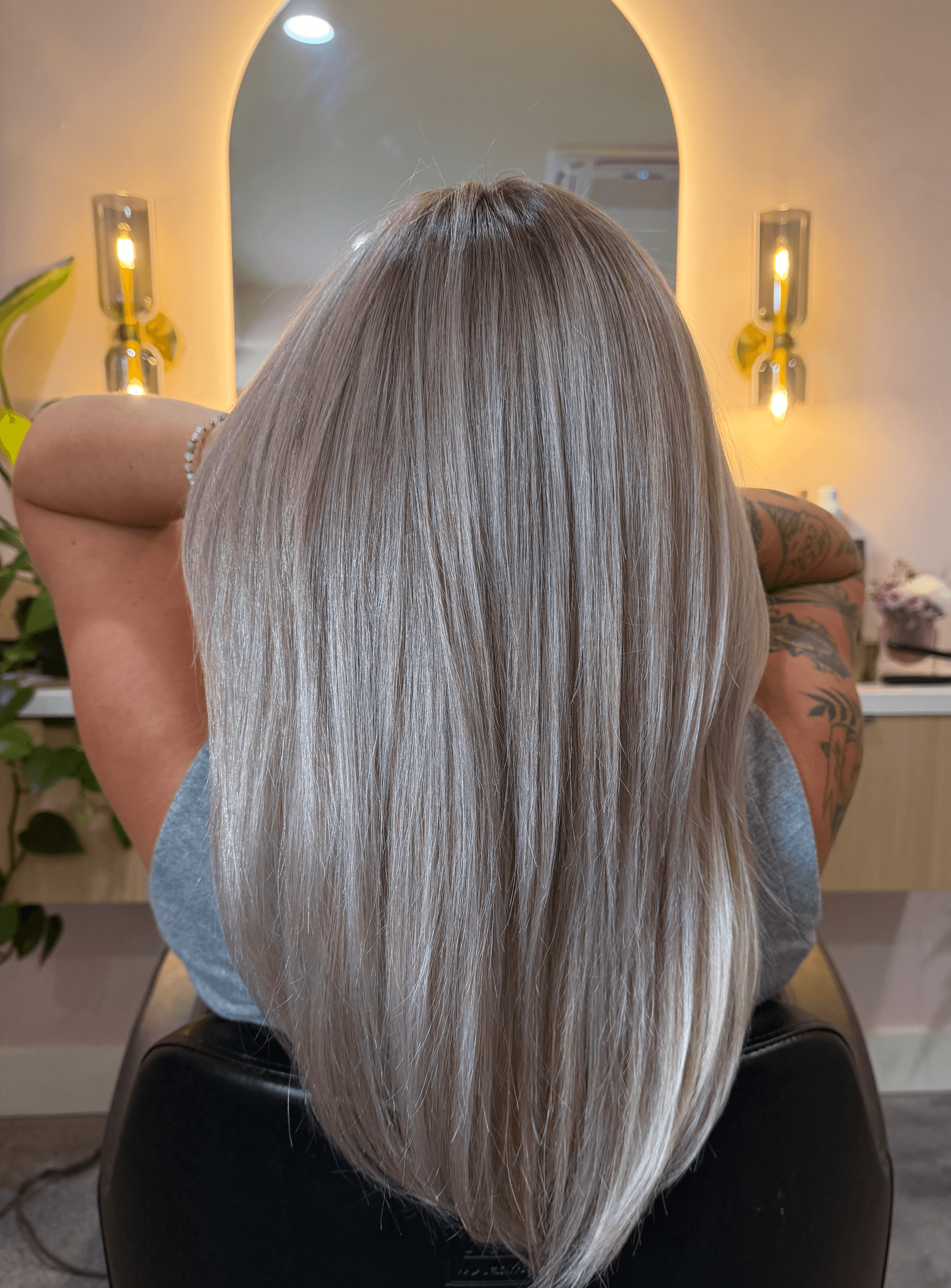 Back of a person with long, straight, silver-blonde hair in a salon, with a mirror and lights in the background.