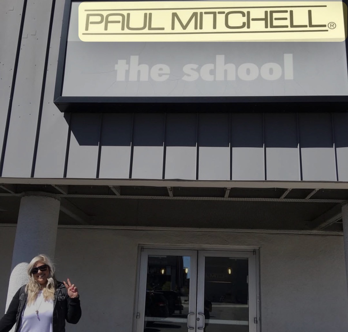 Person with sunglasses and a white shirt standing outside a building with a sign reading 'PAUL MITCHELL' and 'the school'.