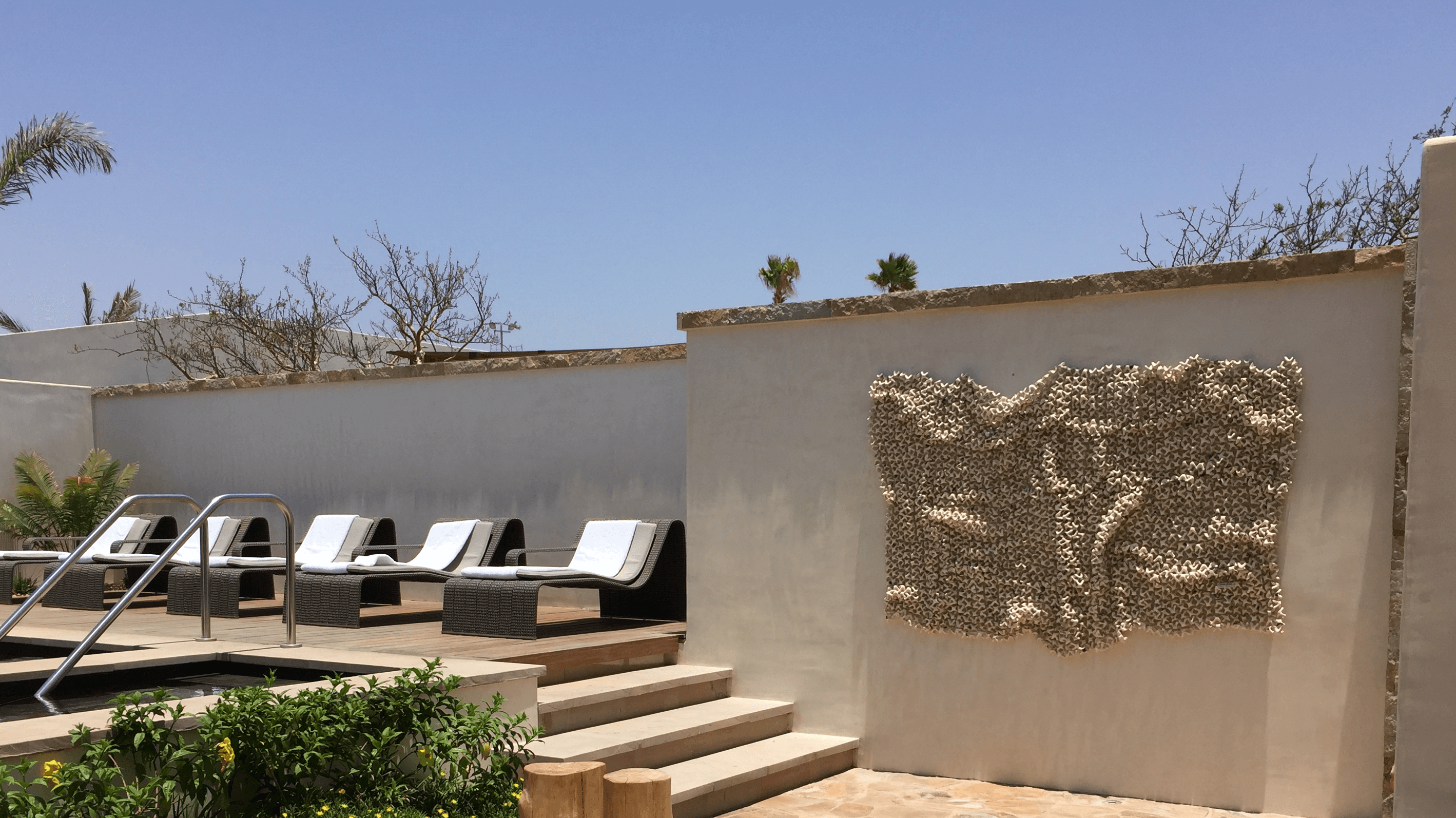 Outdoor area with lounge chairs, a wall with textured artwork, and a clear blue sky.