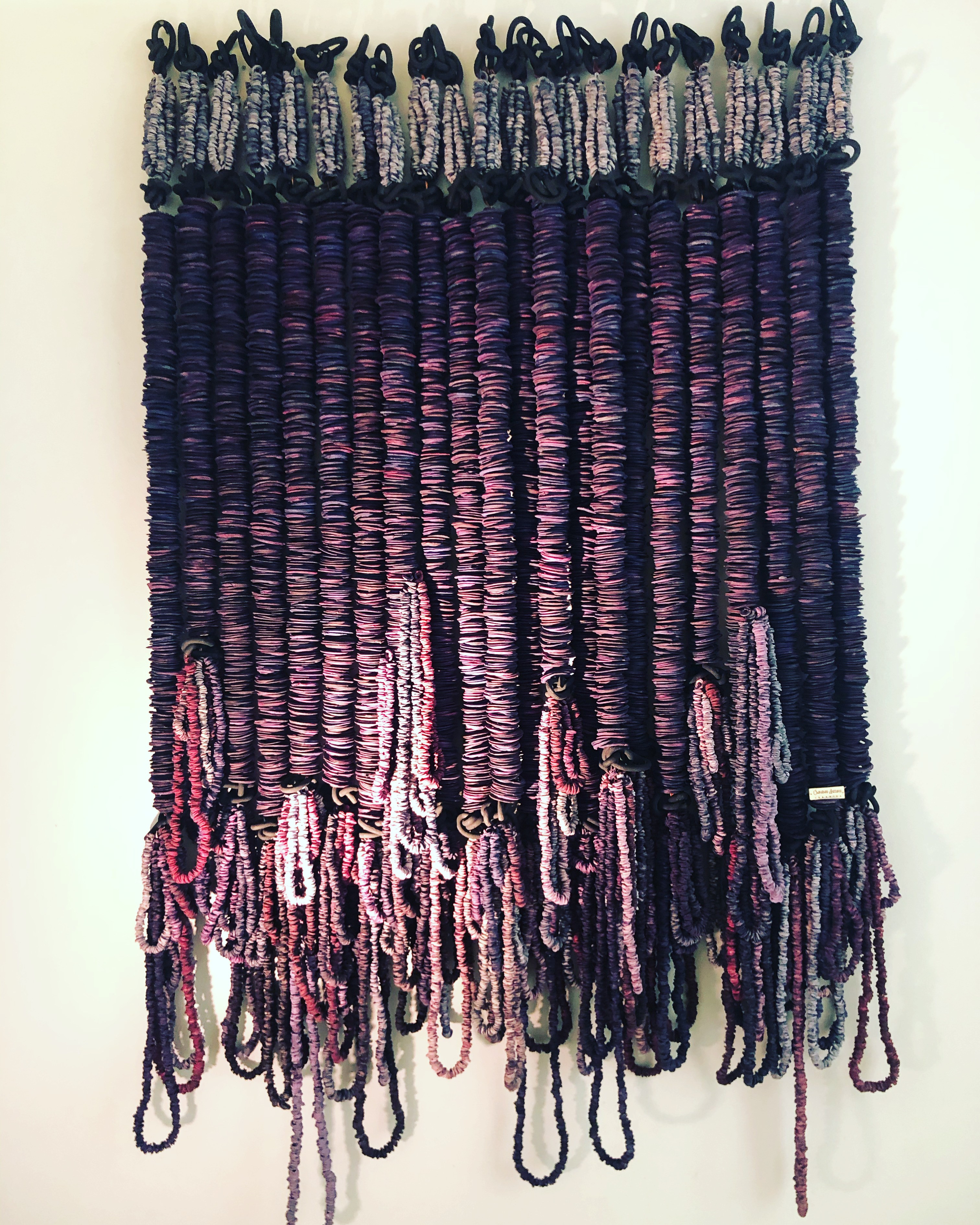 Colorful woven textile with vertical stripes and hanging loops at the bottom, primarily in purple, pink, and black shades.