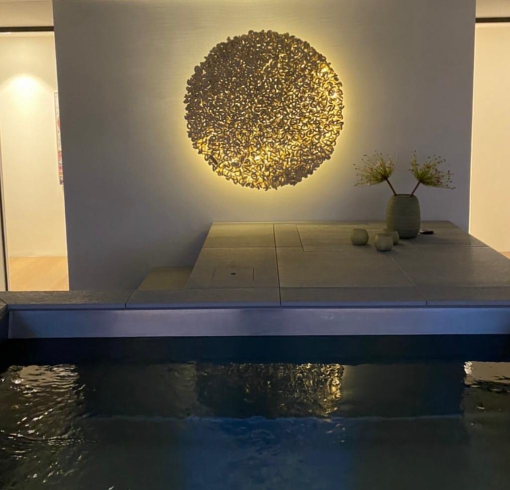 Decorative wall with illuminated circular art piece, vase with flowers, and small objects on table, reflected in water below.