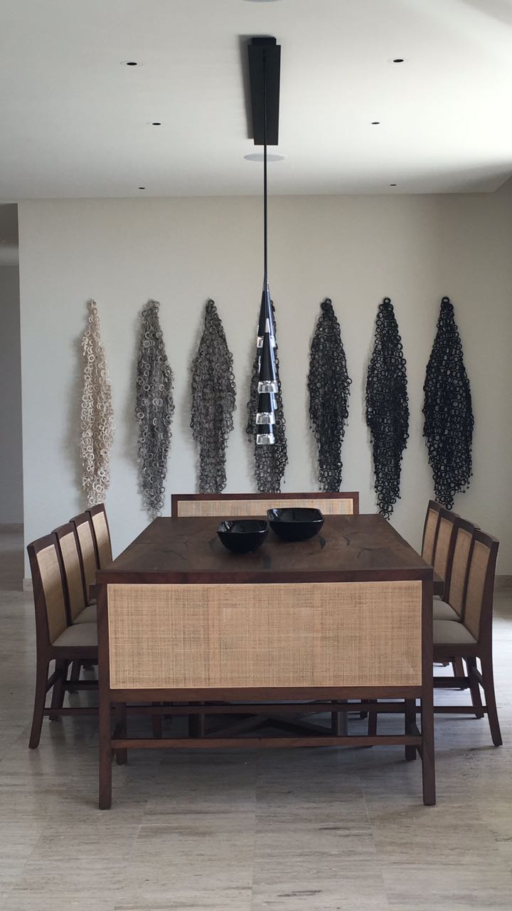Dining table with six chairs in a room with a white wall and tall decorative plants, pendant light hanging above.