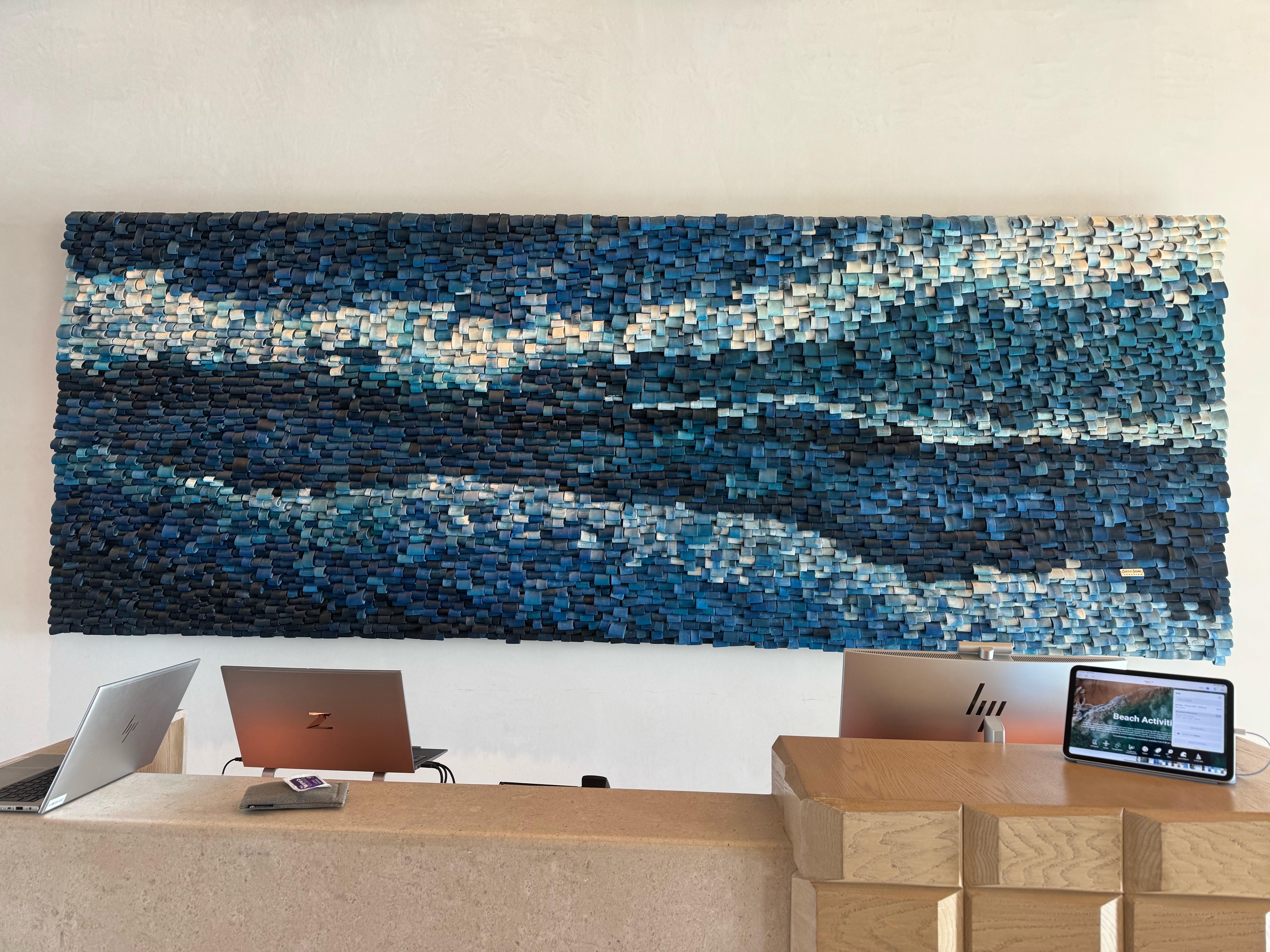 Large mosaic artwork with blue, white, and gray tiles hangs on a white wall above a wooden counter with laptops and a tablet.