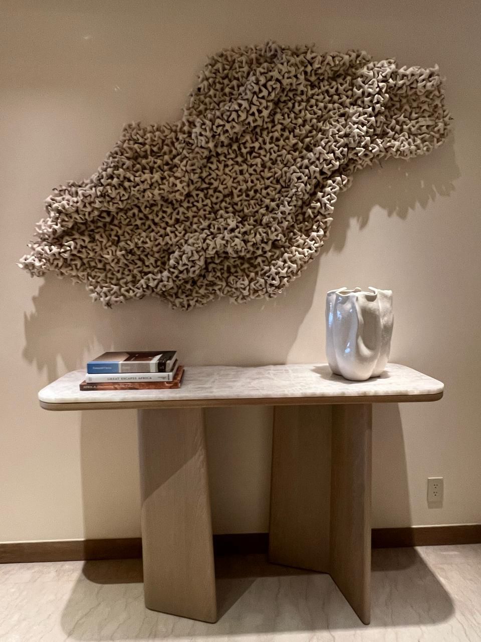 Wooden console table with books and a ceramic vase, wall art above, in a room with beige walls and flooring.