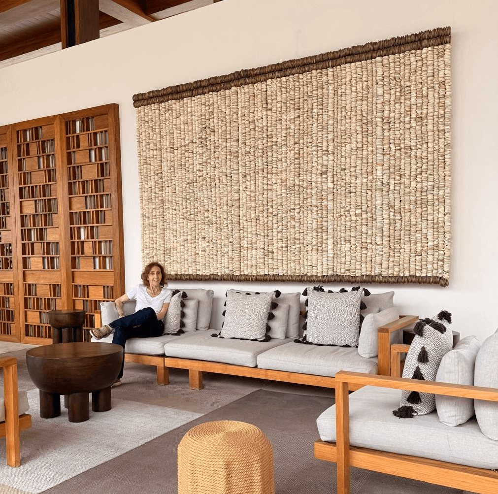 Person sitting on a long sofa with multiple cushions in a room with wooden furniture and a large woven wall hanging.