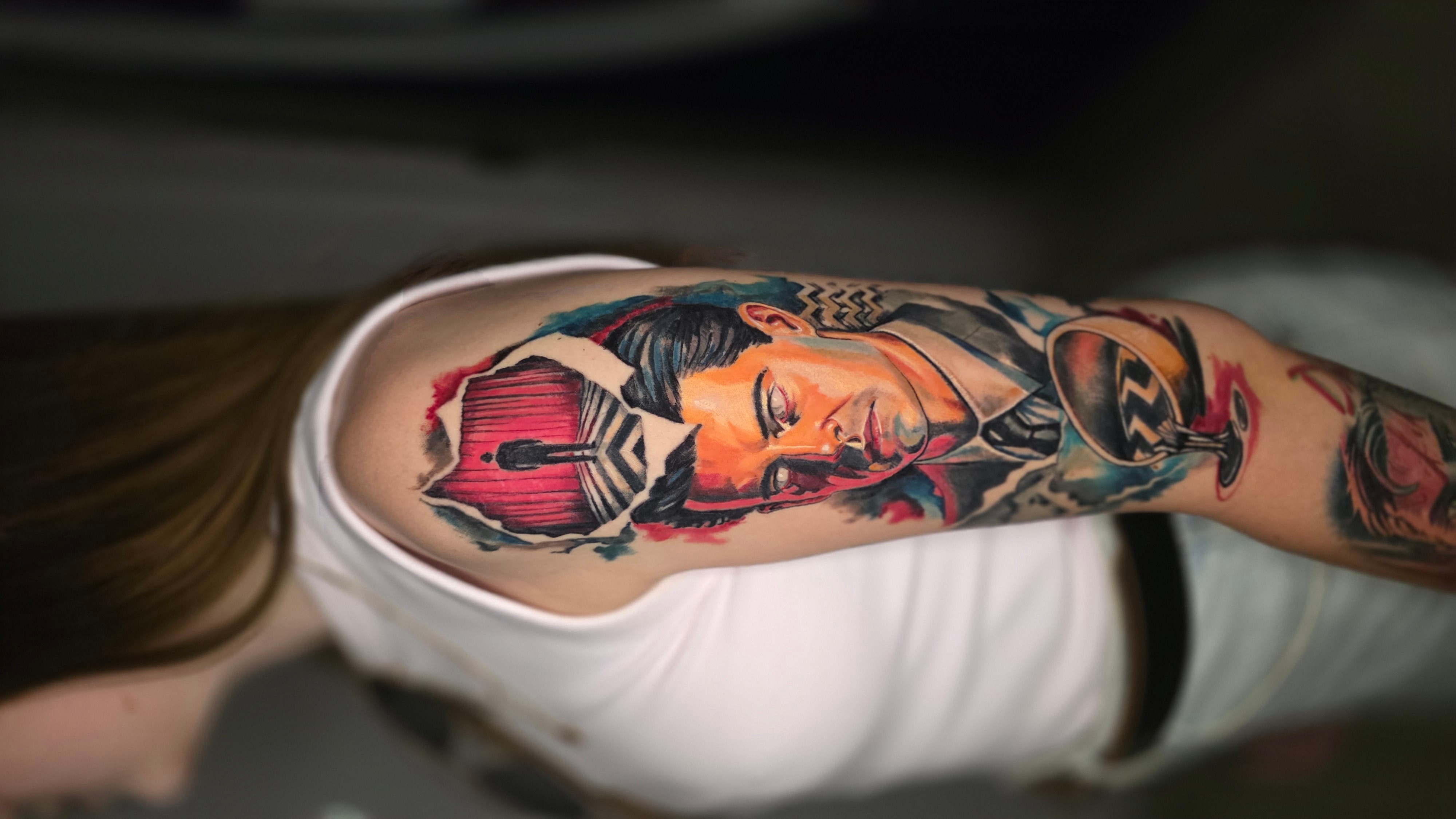 Tattooed arm with a colorful portrait of a woman, skull, and other designs, resting on a white shirt.
