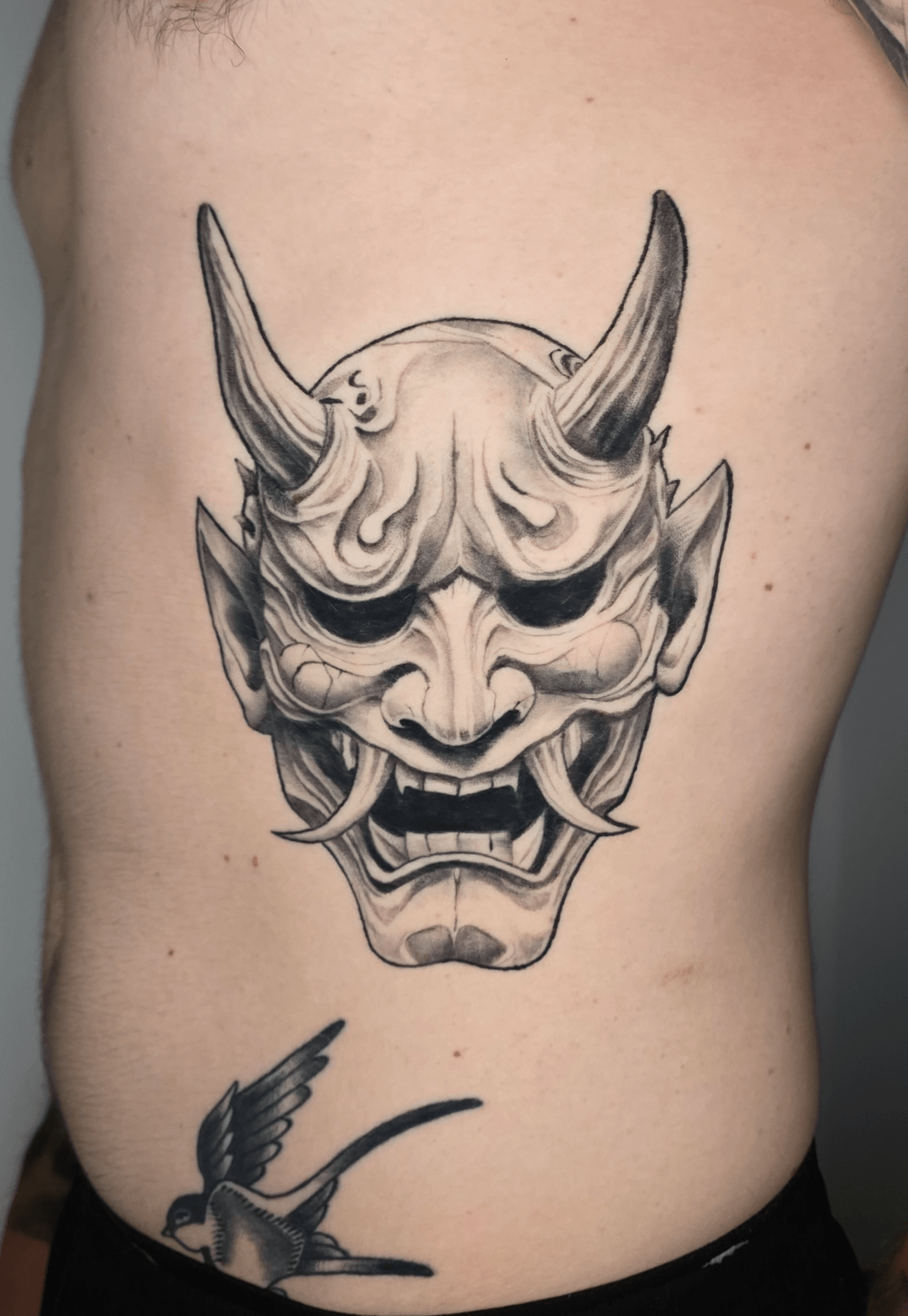 Tattoo of a detailed demon mask with horns, sharp features, and a fierce expression on person's side ribcage.