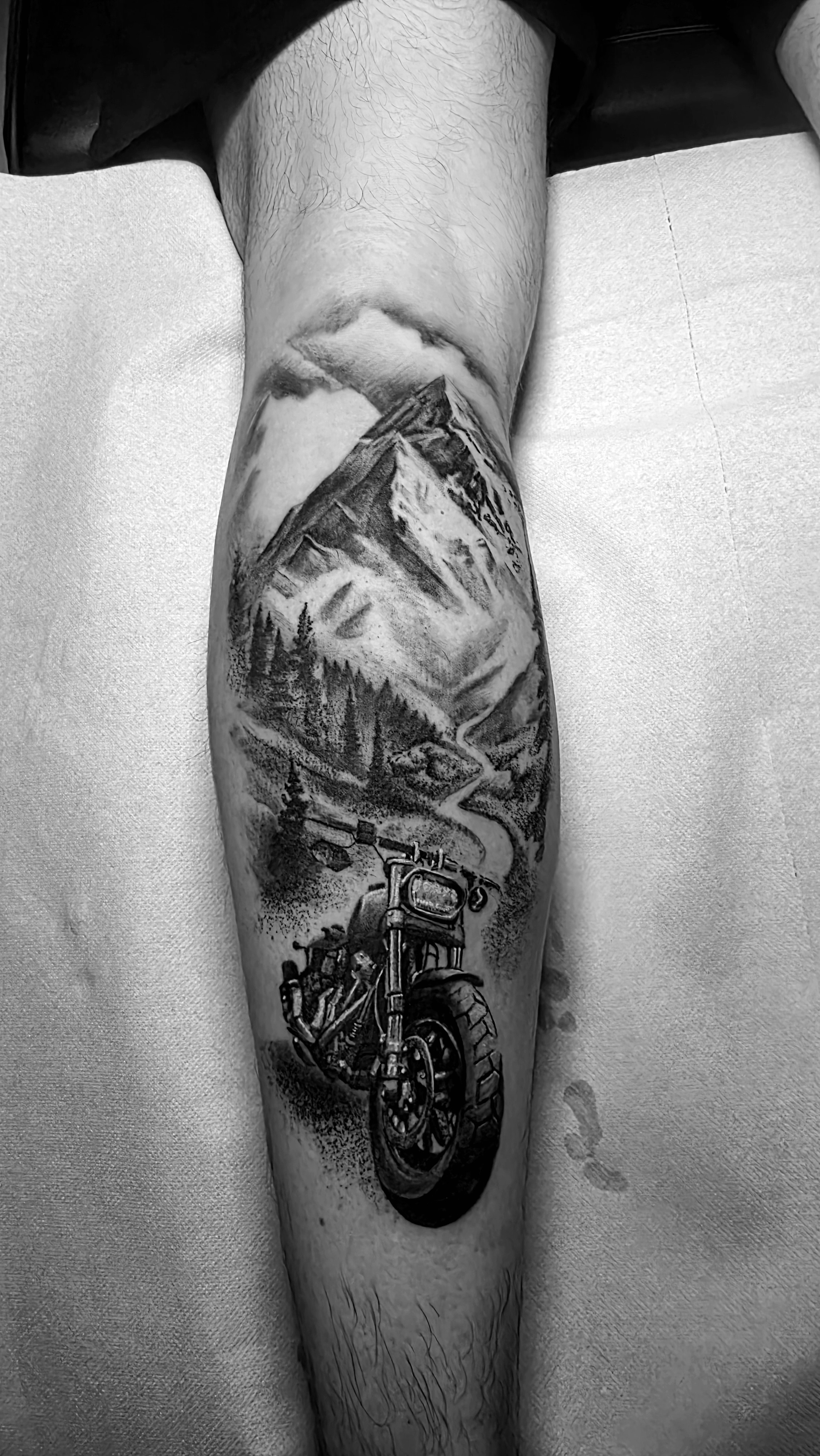 Black and gray tattoo of mountains, trees, a river, and a motorcycle on a person's leg.