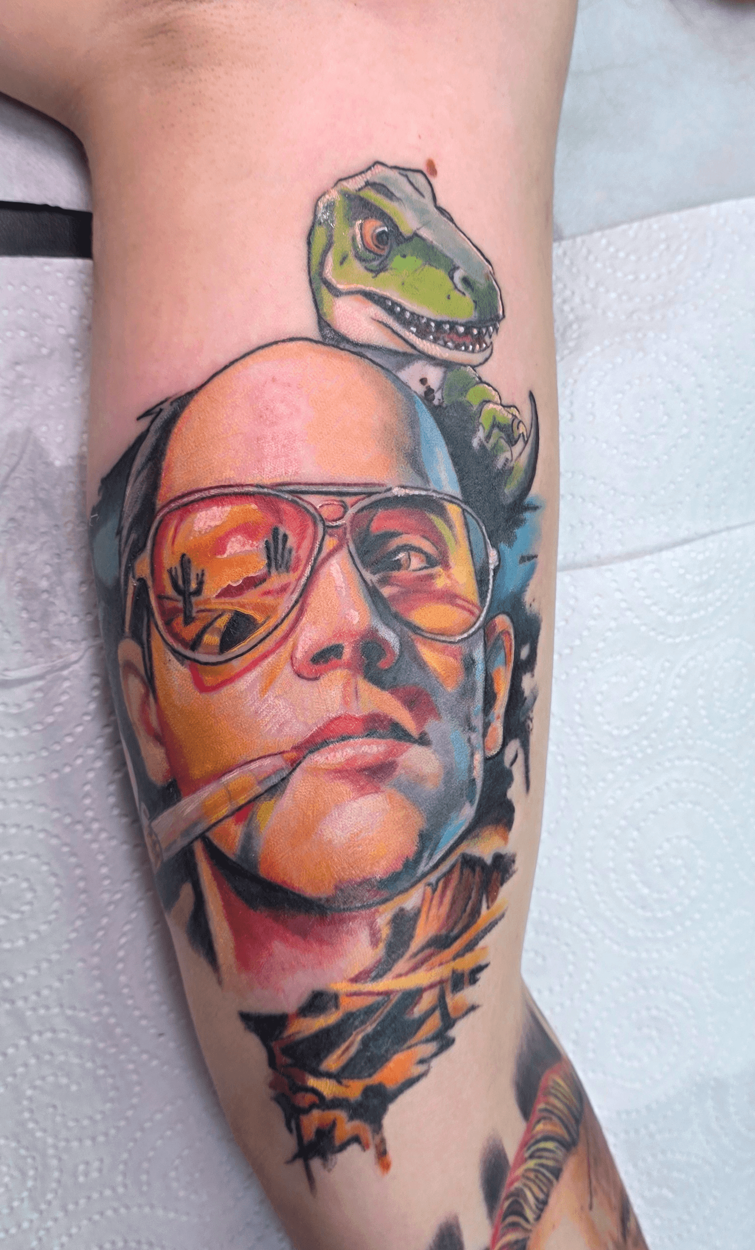Tattoo of a man's face with glasses and a cigar, with a dinosaur head above, on a person's arm.
